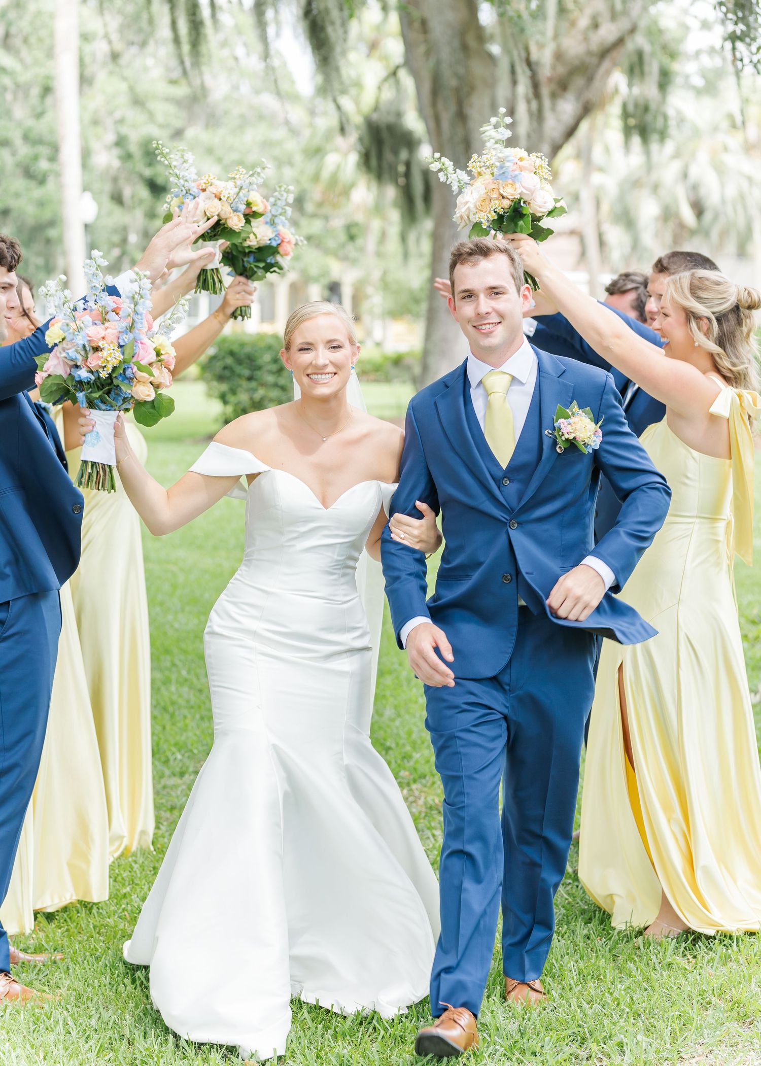 Wedding party in blue suits and yellow dresses poses joyfully on grassy lawn under Spanish moss trees.