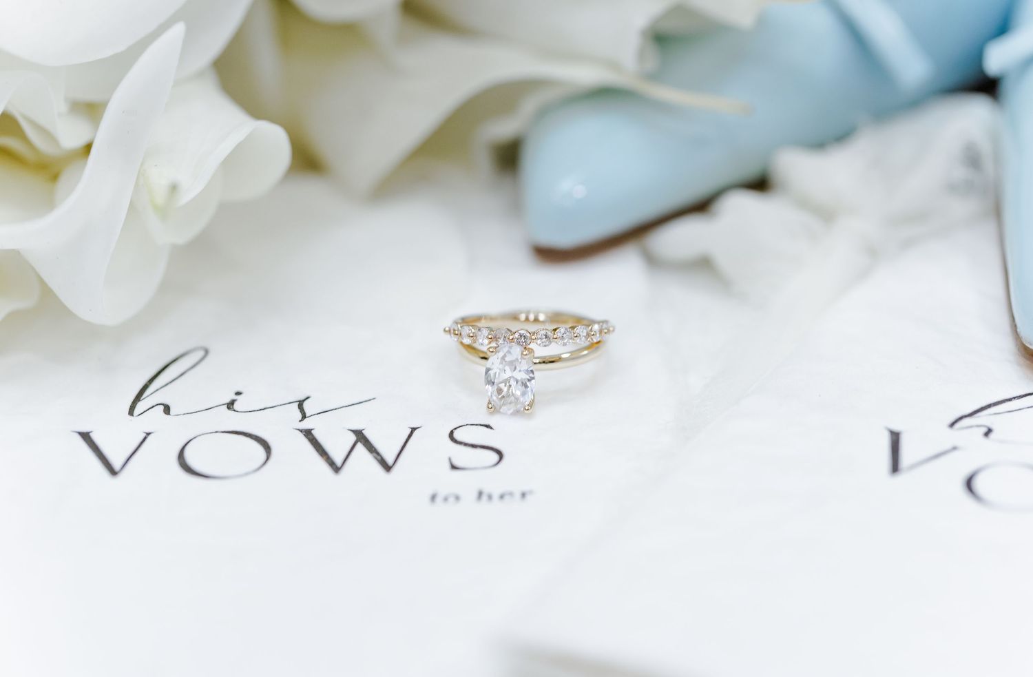 A delicate diamond engagement ring rests on a white paper with 'VOWS' text and soft blue floral decor.
