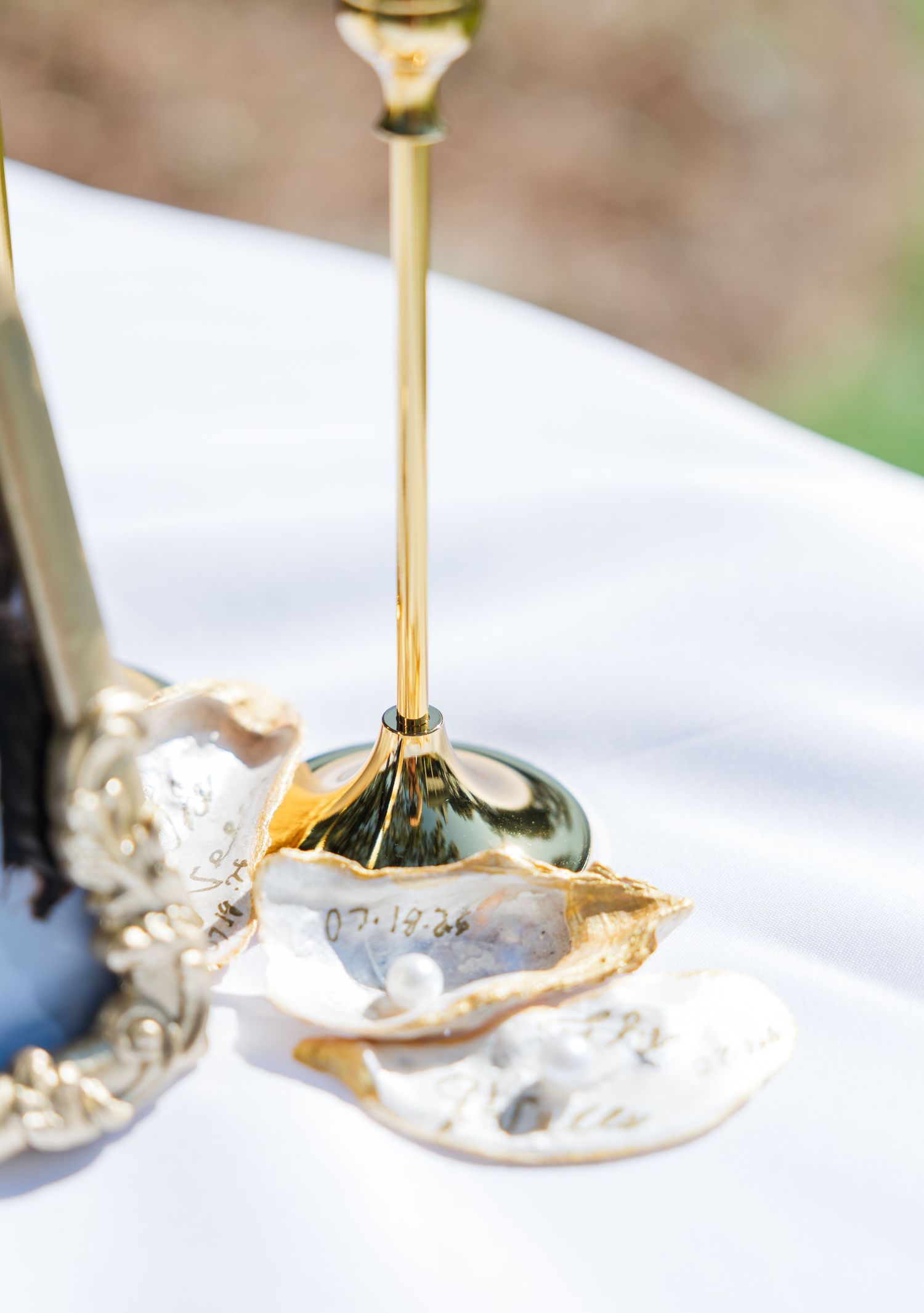 Gold candlestick holder beside an oyster shell with pearls and a wedding date on a white tablecloth.