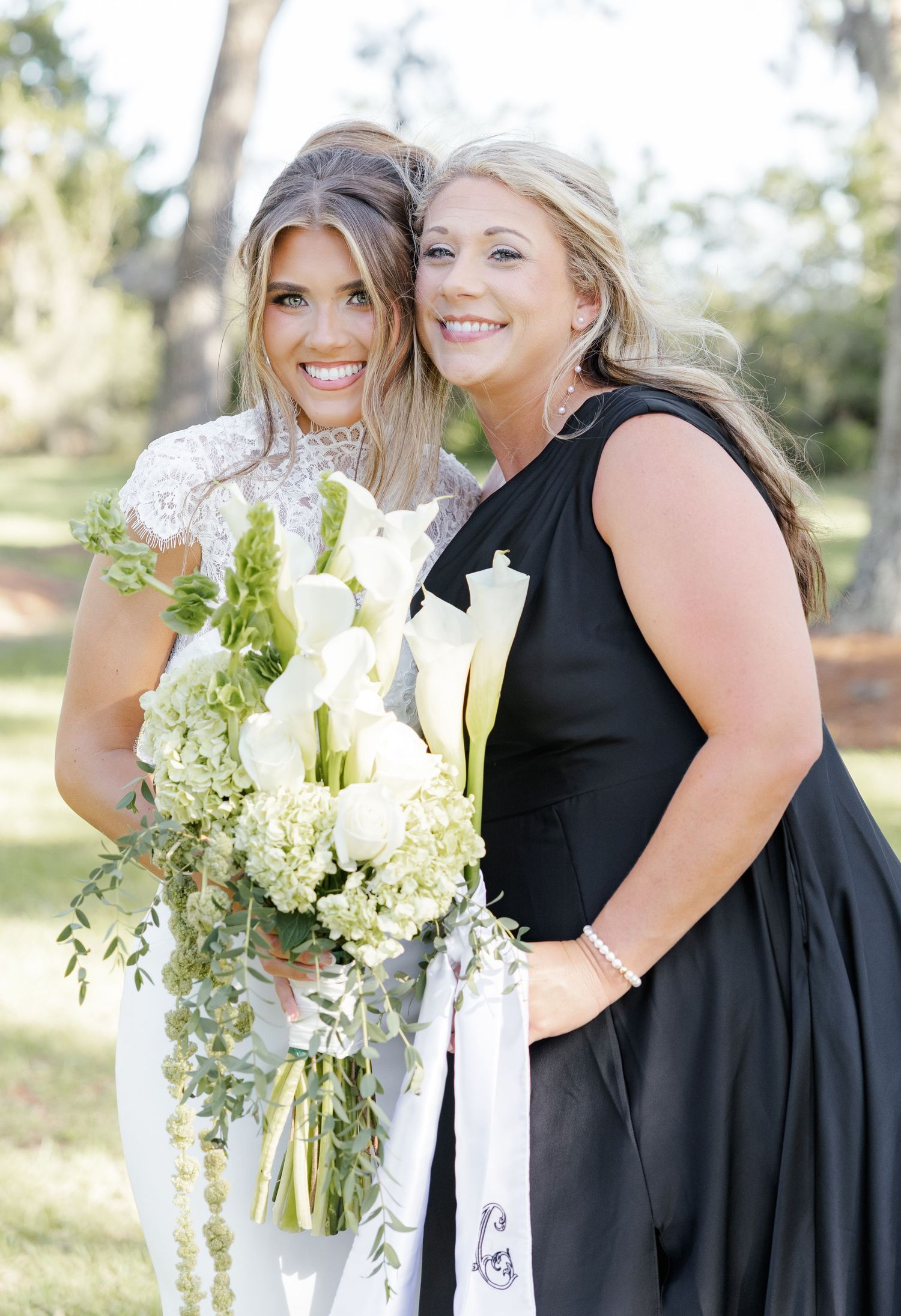 Bride in white lace gown holding white calla lily bouquet poses with bridesmaid in black dress outdoors.