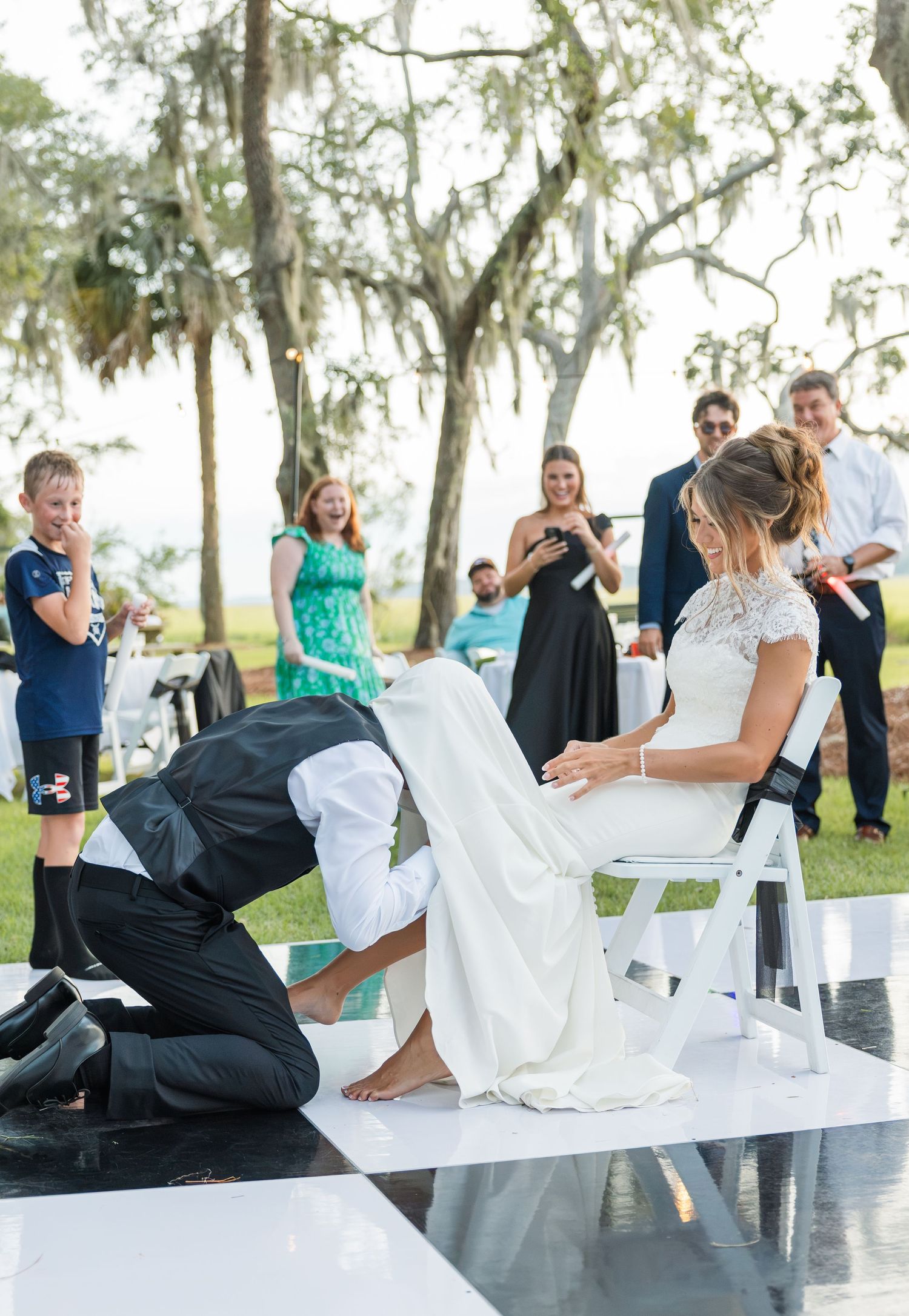Groom removes bride's garter at outdoor wedding reception on black and white dance floor.