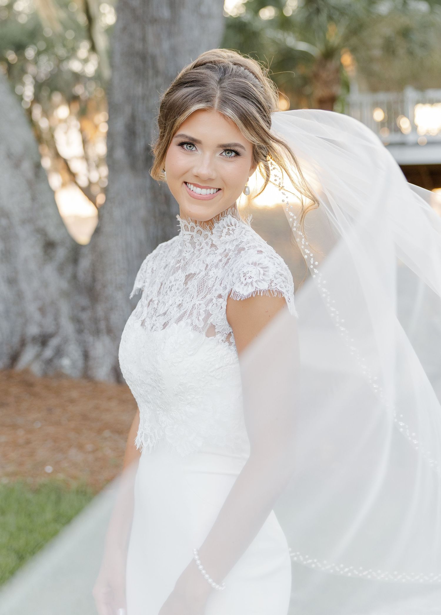 Smiling bride in lace high-neck wedding gown with flowing veil outdoors among trees in soft natural light.