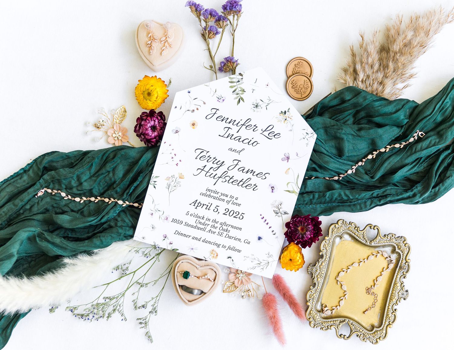 Elegant wedding invitation with emerald green velvet fabric, dried flowers, and gold accents arranged on white background.