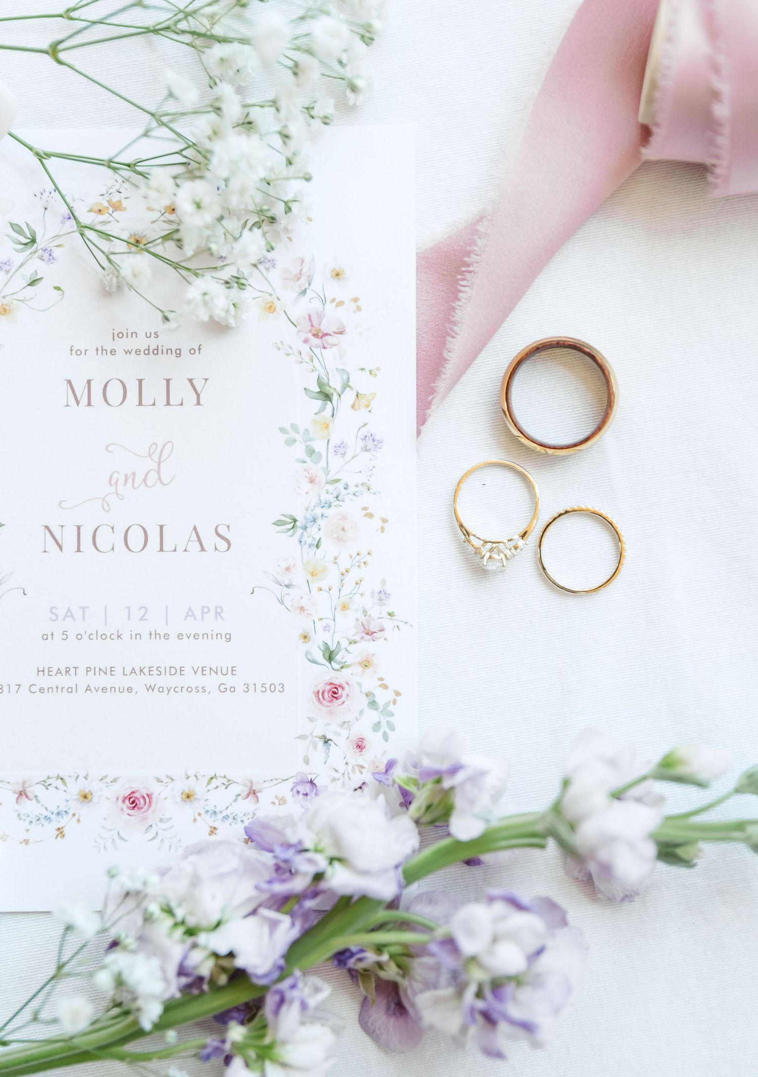 Romantic wedding stationery flatlay featuring rings, pastel ribbons, and soft purple flowers with invitation card.