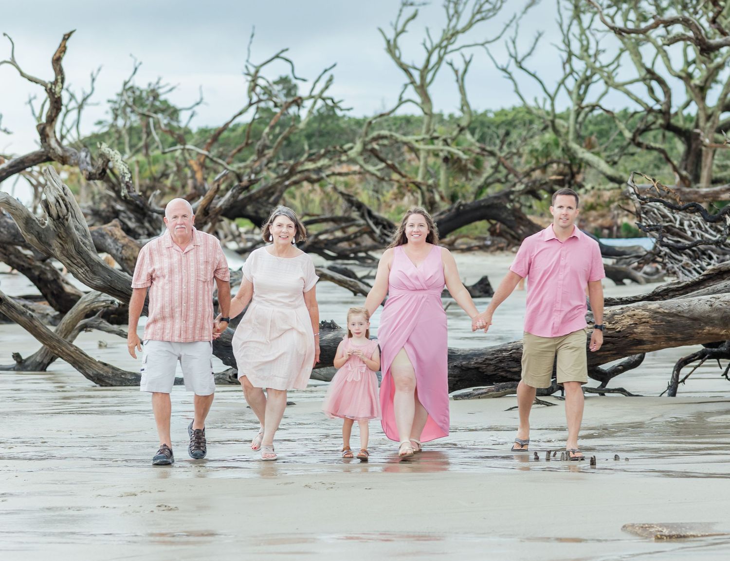 Six people wearing pink and white outfits sit casually along a large driftwood tree trunk at the beach.