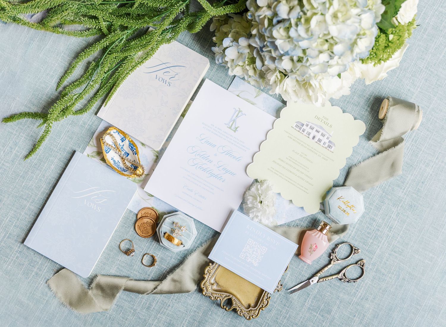 Elegant wedding stationery flat lay with floral accents, ribbons, rings, and scissors on a soft blue backdrop.