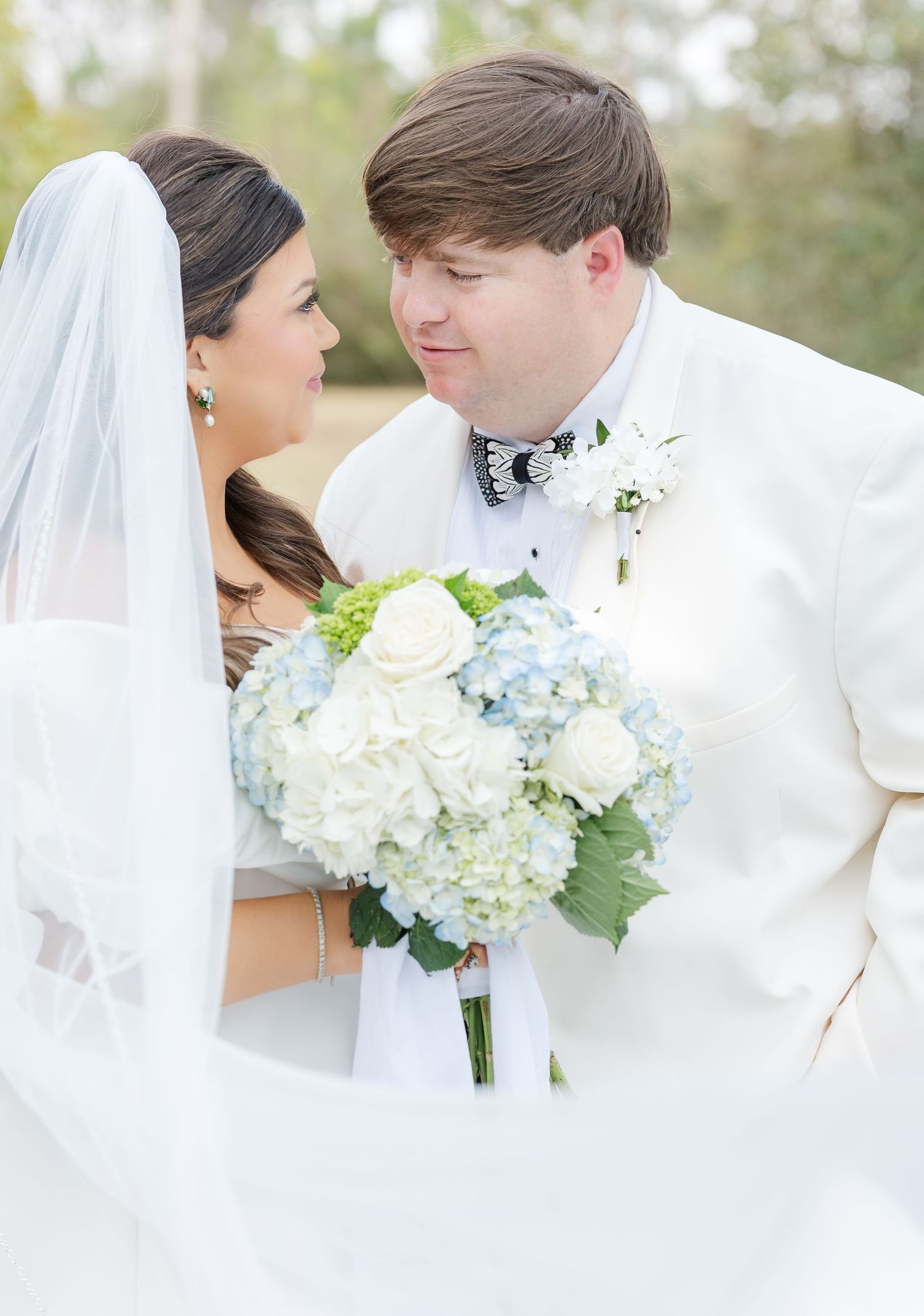 Bride holds blue hydrangea and white rose bouquet while groom in white tuxedo smiles at her.