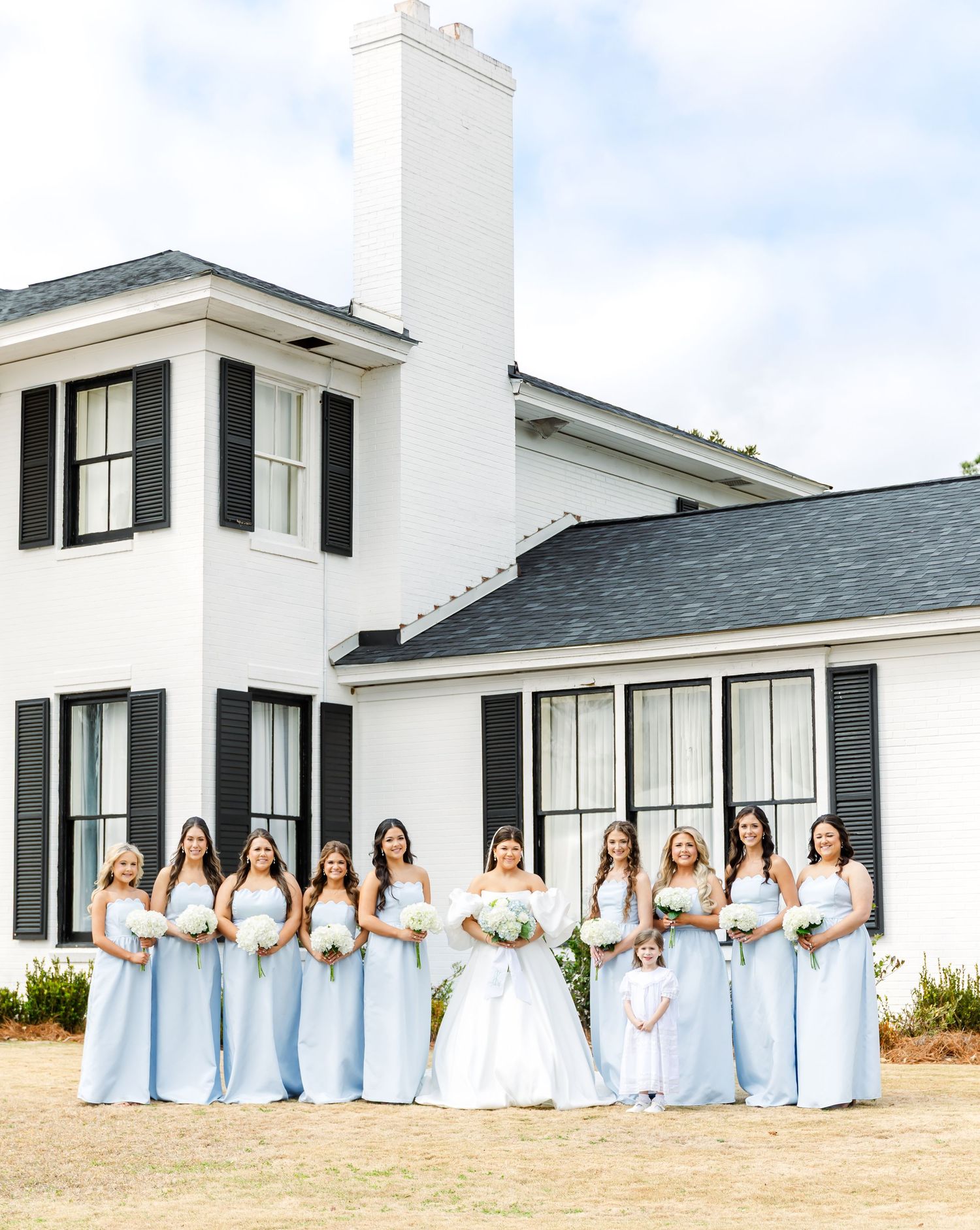 Bride with bridesmaids in light blue gowns holding white bouquets outside elegant white venue.