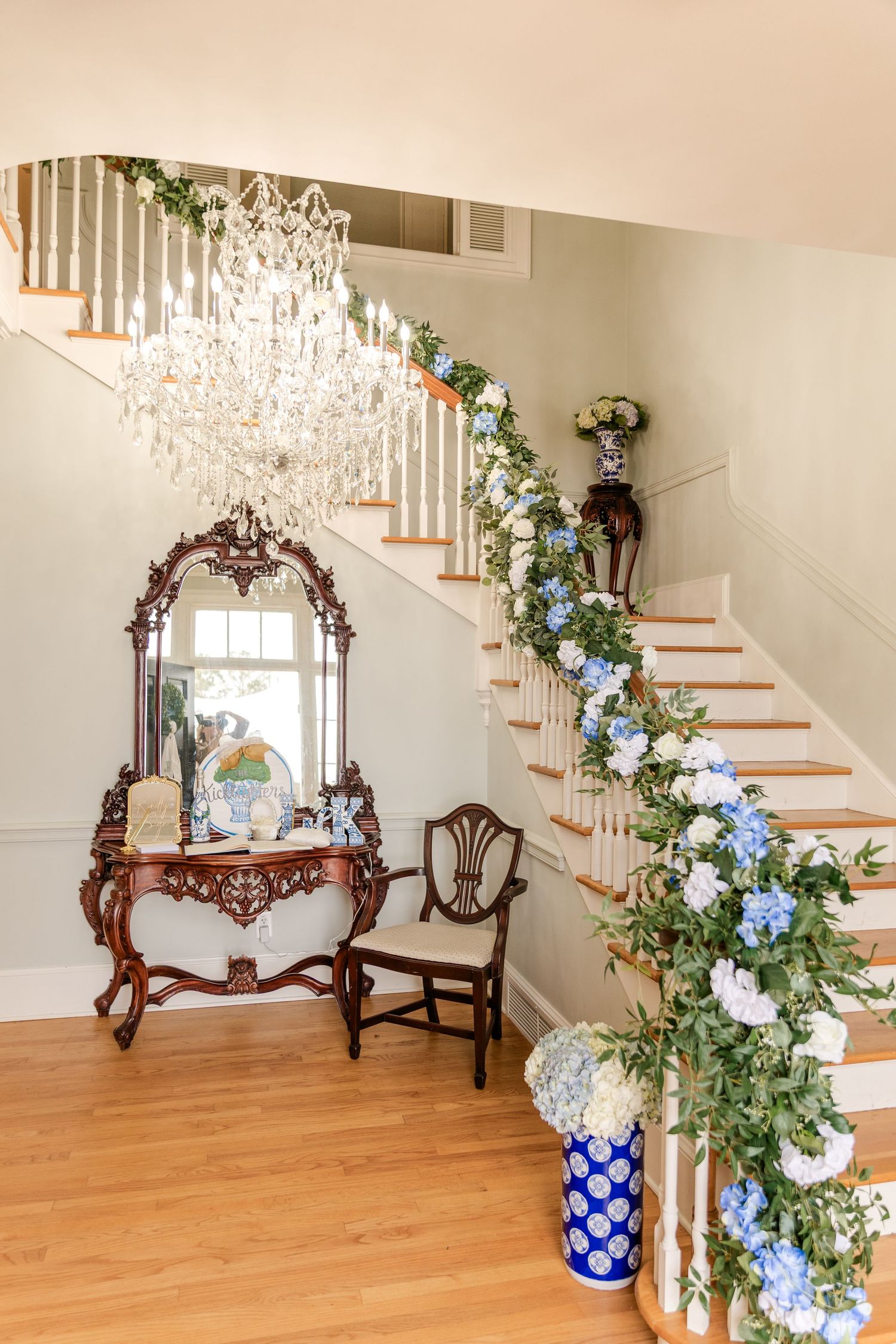 Grand staircase adorned with blue and white hydrangea garland beneath a crystal chandelier in wedding venue.