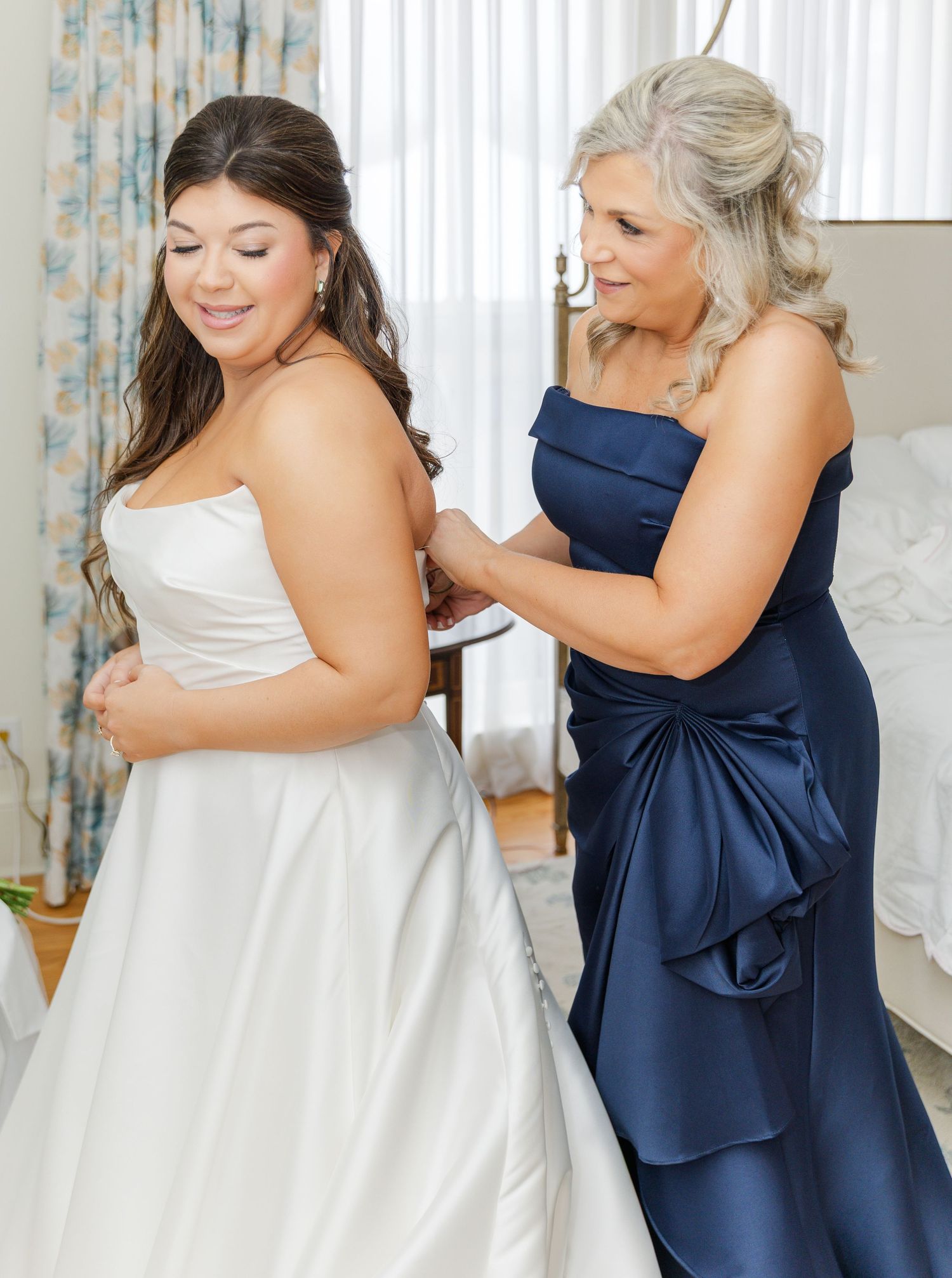 Bride in white strapless gown gets dress fastened by woman in navy blue satin gown during wedding preparations.