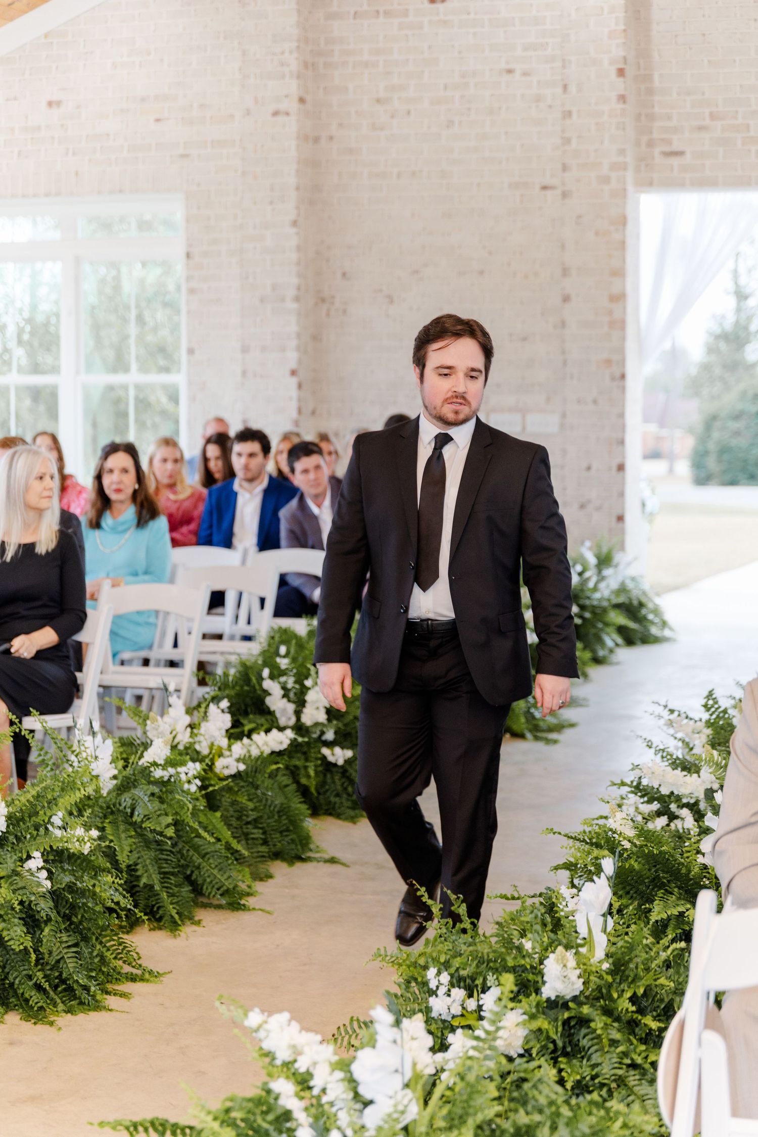 Groomsman in black suit walks down floral-lined aisle in bright white brick ceremony venue.