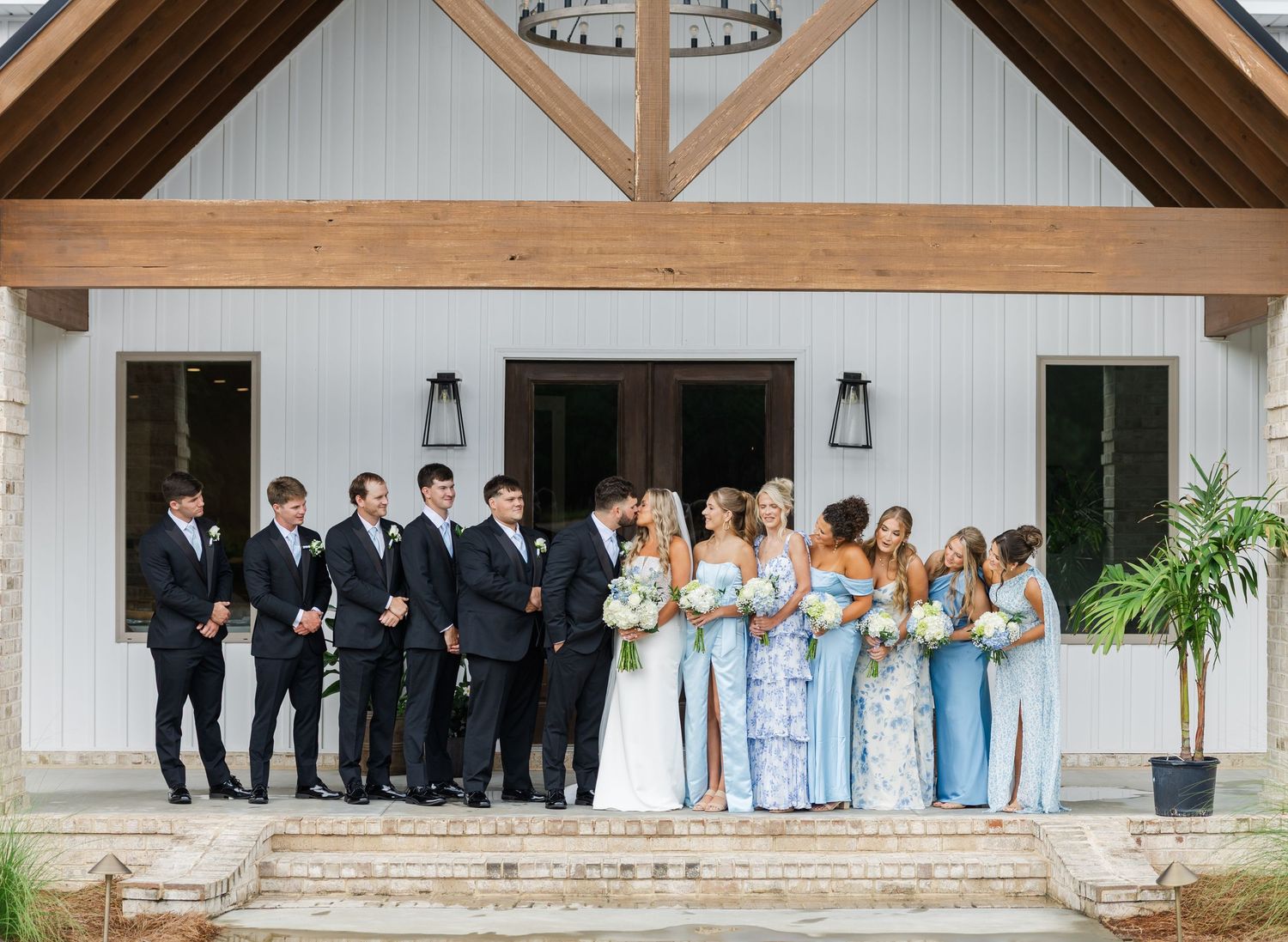 Full wedding party gathered on steps of white barn venue with wood beam entrance.