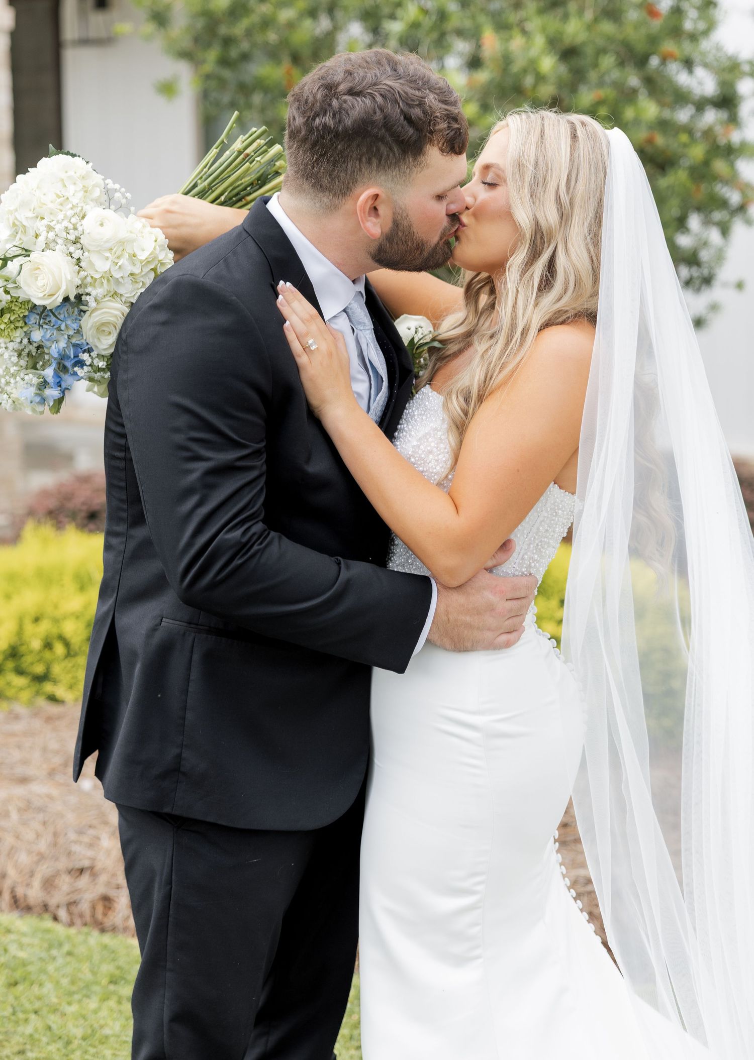 Bride and groom share a romantic kiss outdoors, bride holding blue and white bouquet wearing lace gown with veil.