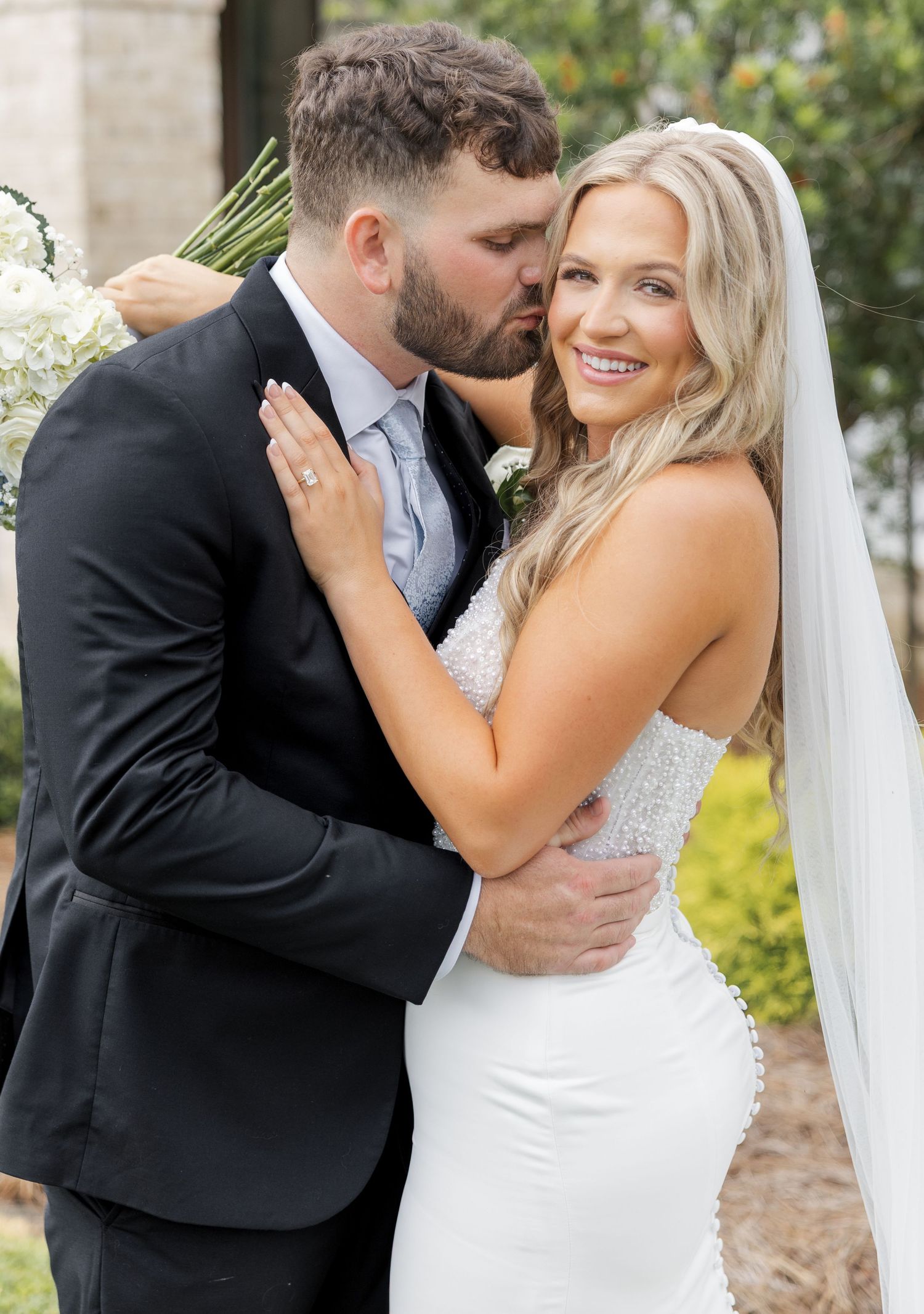 A groom in a black suit kisses his smiling bride in a white gown and veil on their wedding day.