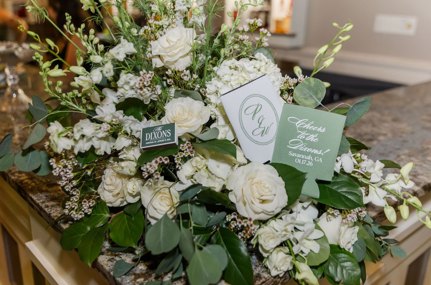 Lush white rose and greenery floral arrangement with monogram cards on marble surface.