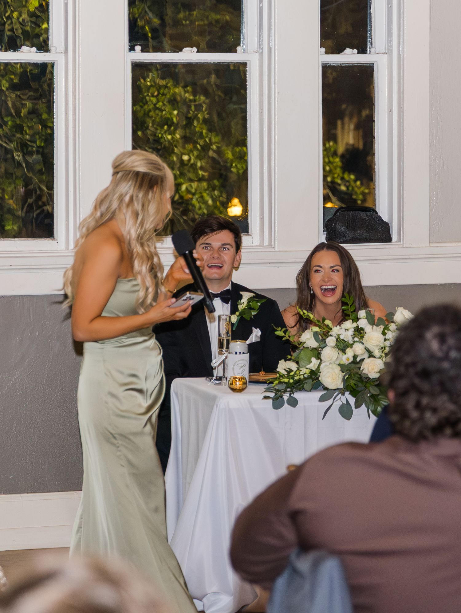 A bridesmaid gives a toast as the bride and groom laugh joyfully at their wedding reception table.