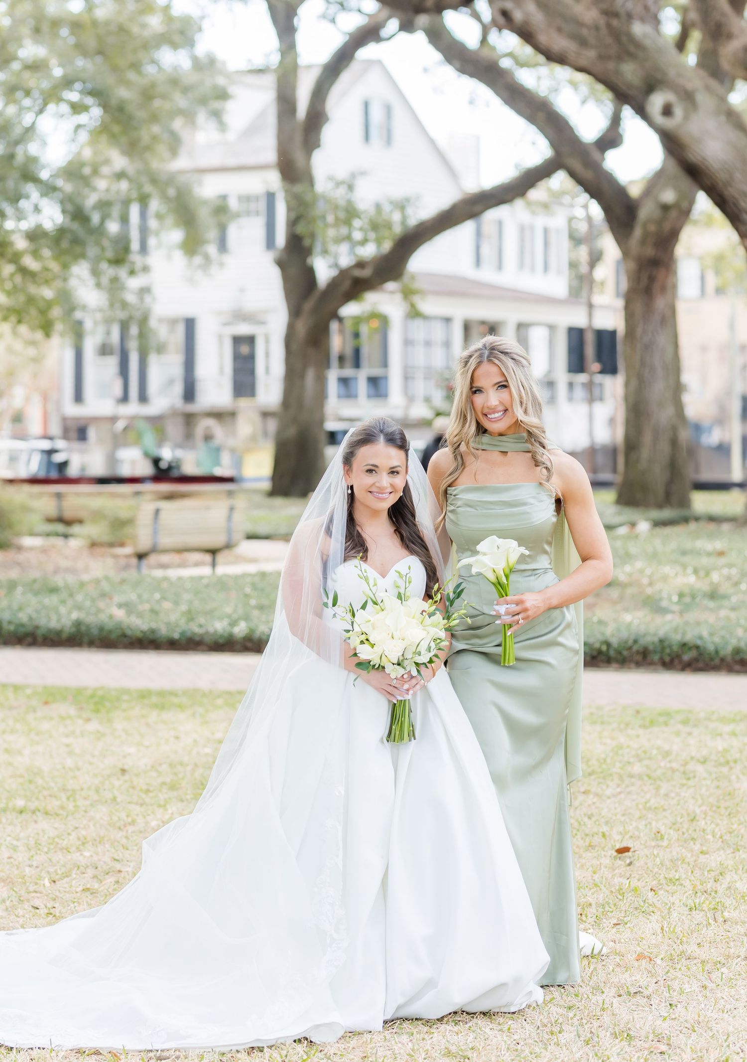 Bride in white ball gown with veil stands beside bridesmaid in sage green satin dress holding white bouquets outdoors.