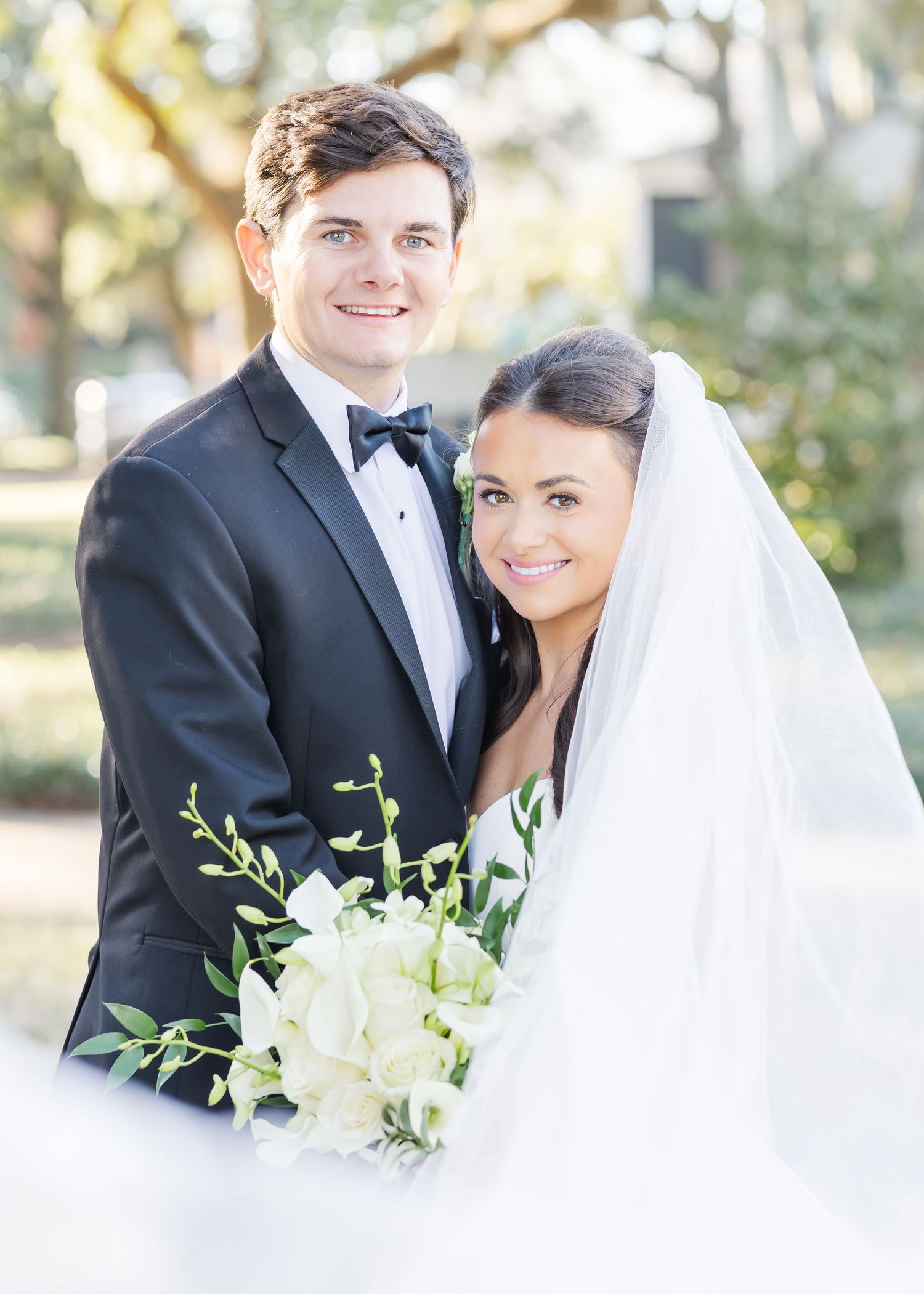 Bride and groom portrait outdoors, she holds white orchid bouquet, both smiling at camera.