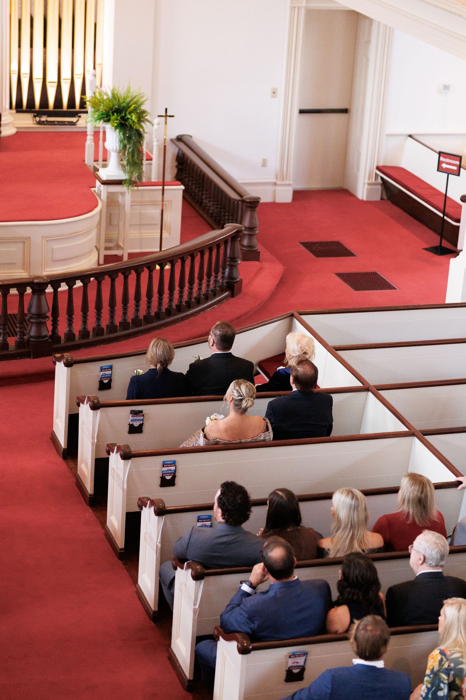 Aerial view of wedding guests seated in white church pews with red carpet aisle during ceremony.