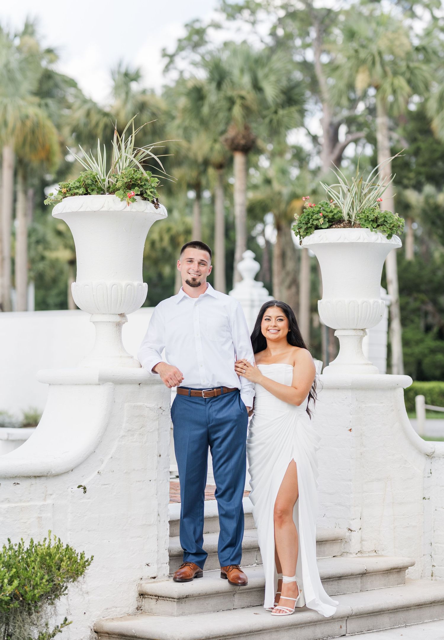 Couple posing on white stone steps with large decorative urns and palm trees during an outdoor engagement session.