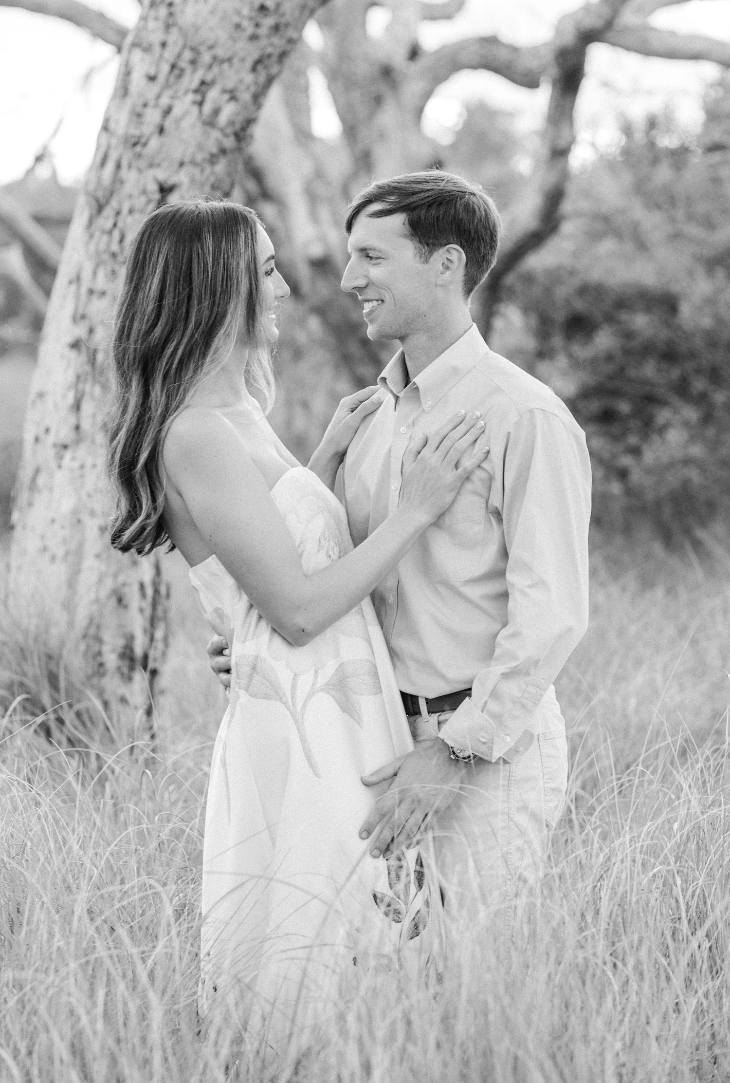 Black and white photo of couple facing each other in tall grass field, woman in white dress, man in button-up shirt.