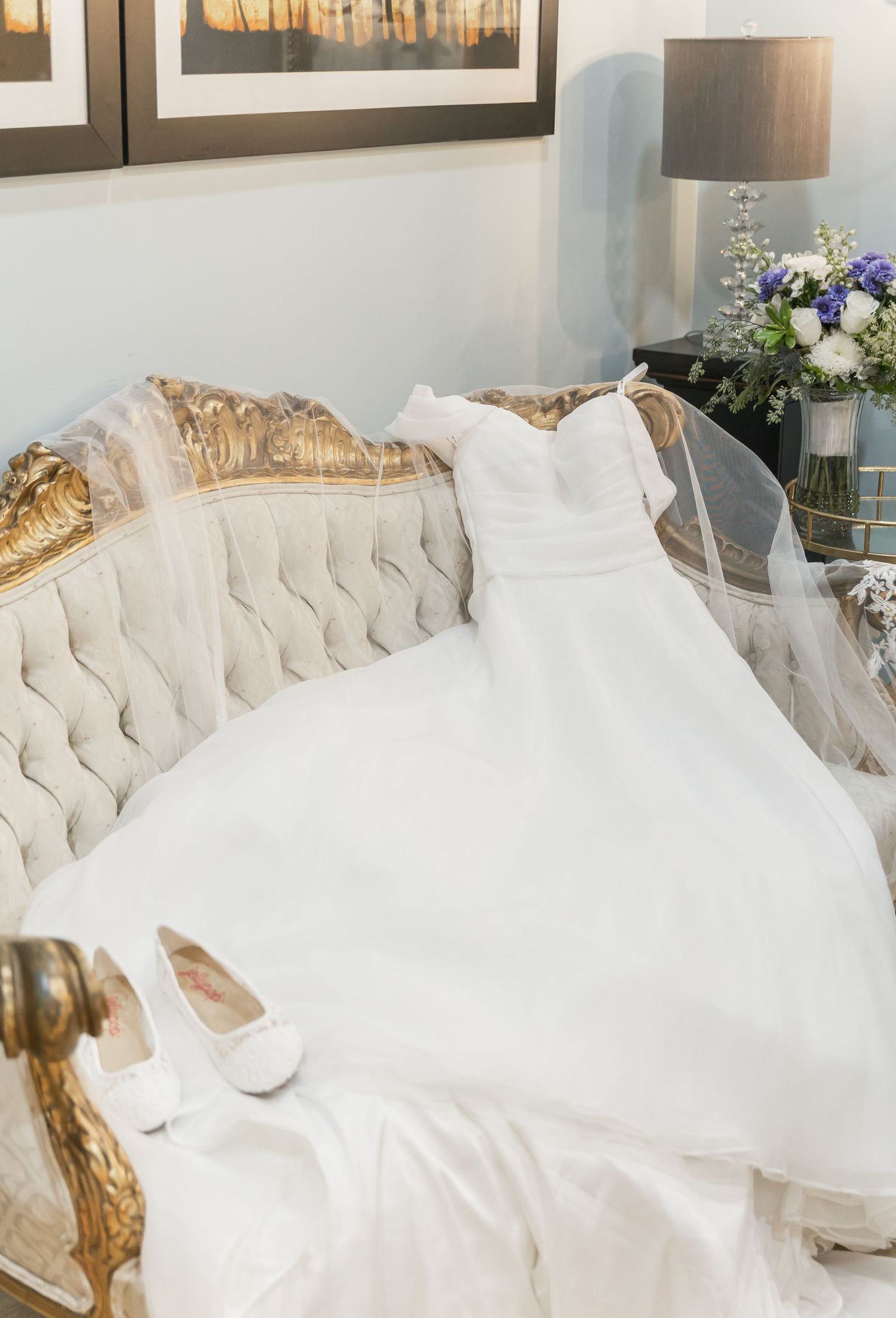 An elegant white wedding dress and veil displayed on a tufted cream sofa with white shoes and floral decor.