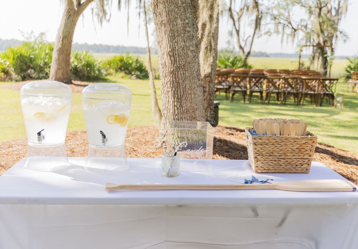 A white-clothed table setup outdoors with empty glasses and a wicker basket near a wooden bridge and waterfront view.