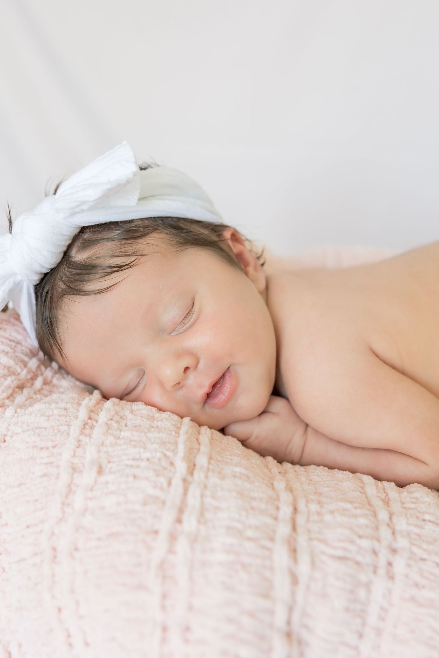 Sleeping newborn with white bow headband rests on pink textured fabric.