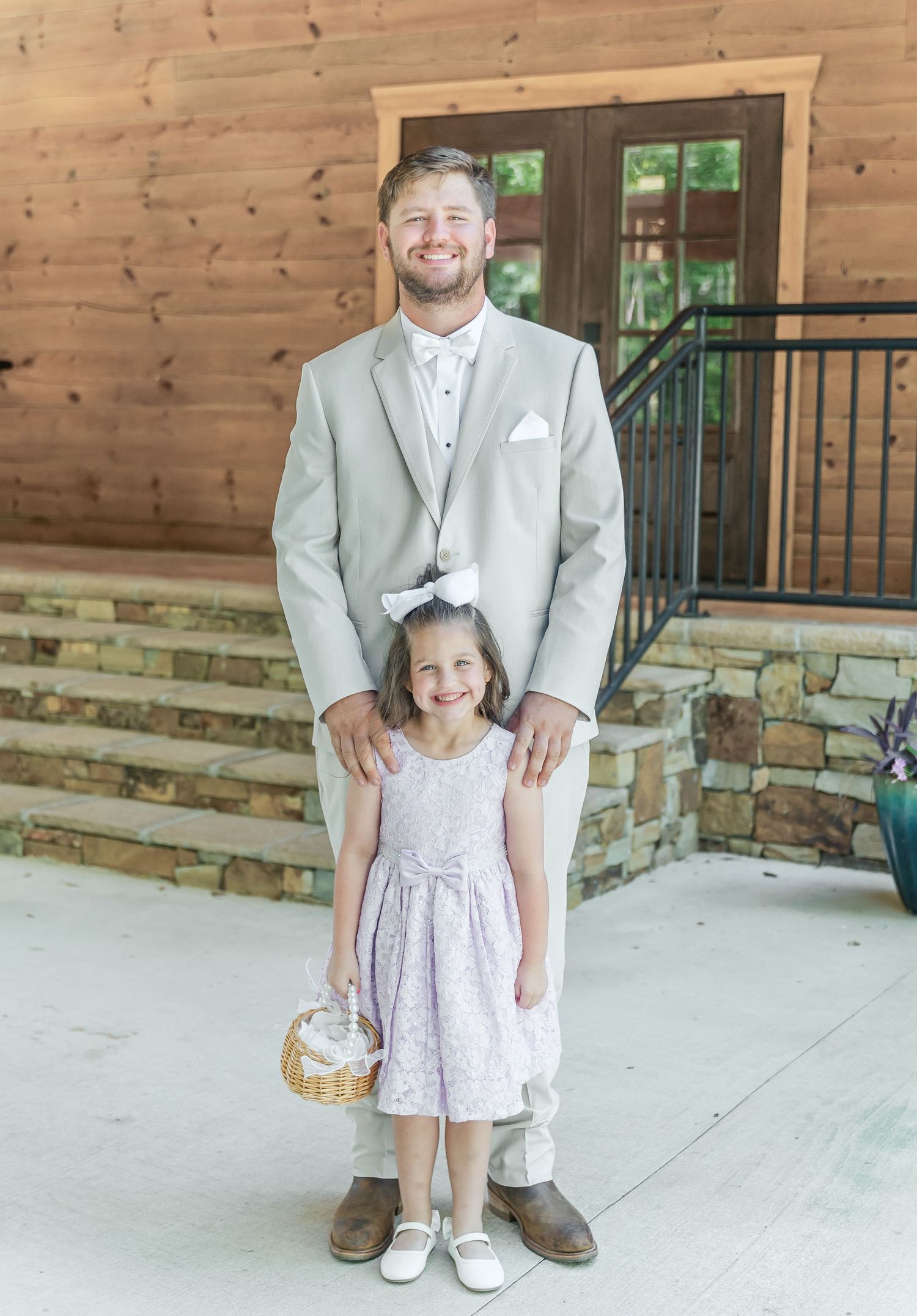 Person in light suit stands with young flower girl in pink dress on wooden porch.