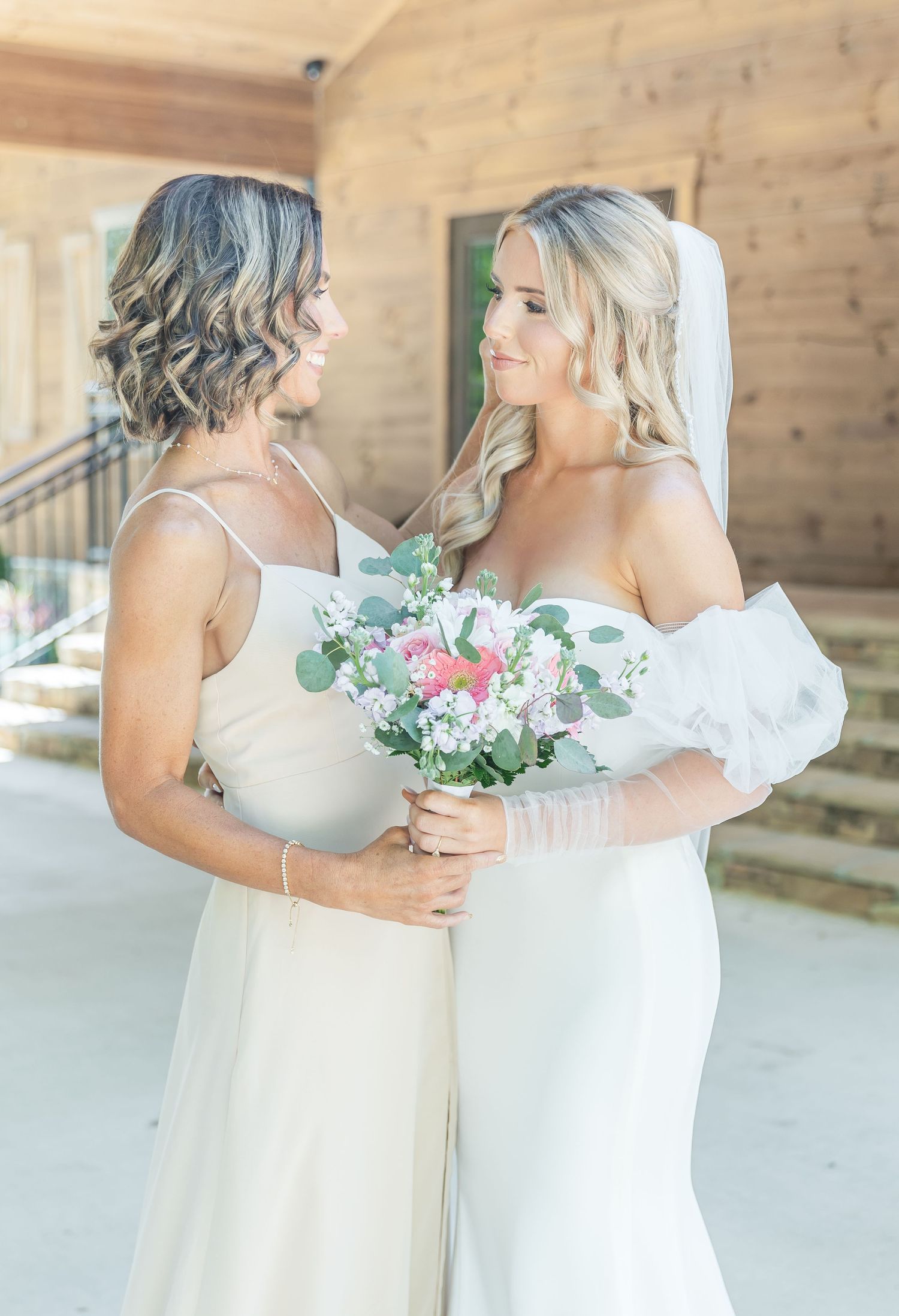 Bride and bridesmaid wearing white dresses share a tender moment while holding a floral bouquet.