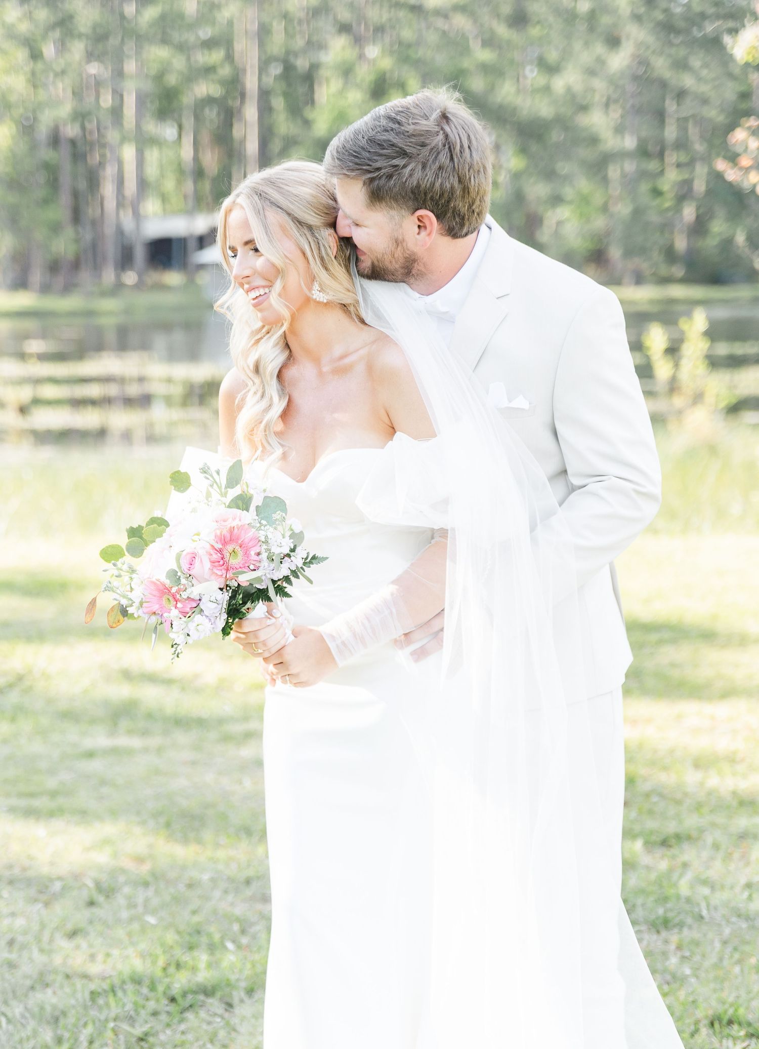 Romantic wedding couple shares intimate moment with pink bouquet against wooded backdrop.