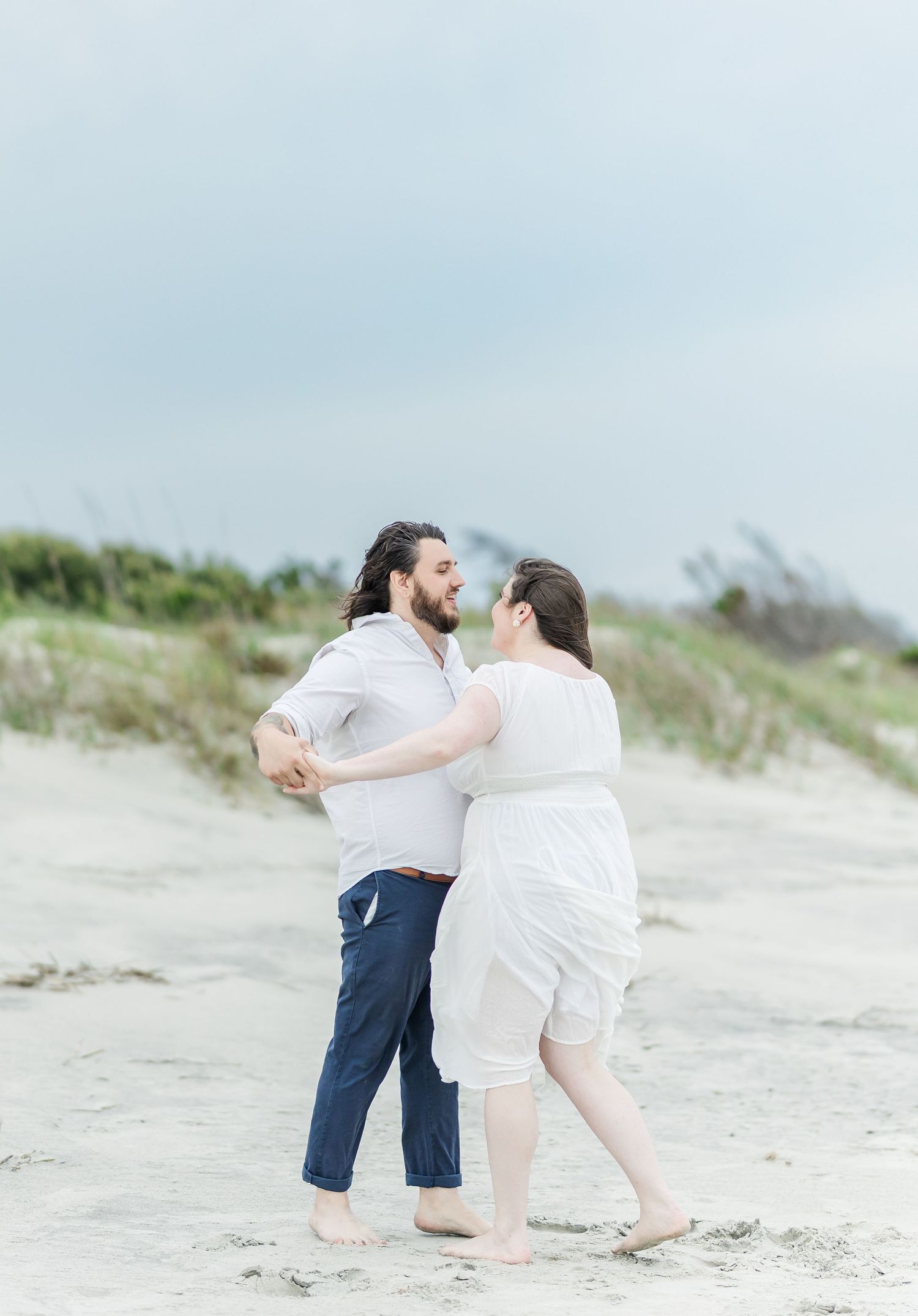A couple embraces while dancing on a sandy beach with dunes in the background.