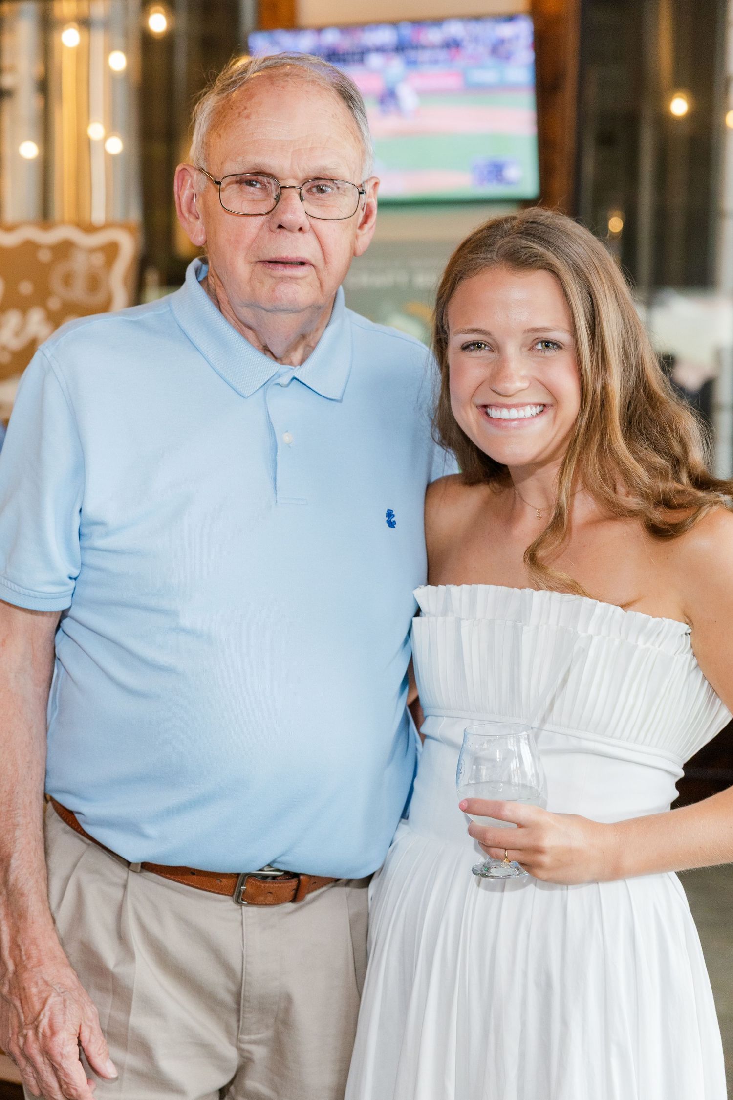 Elderly man in blue polo and young woman in white strapless dress pose together at an engagement party.