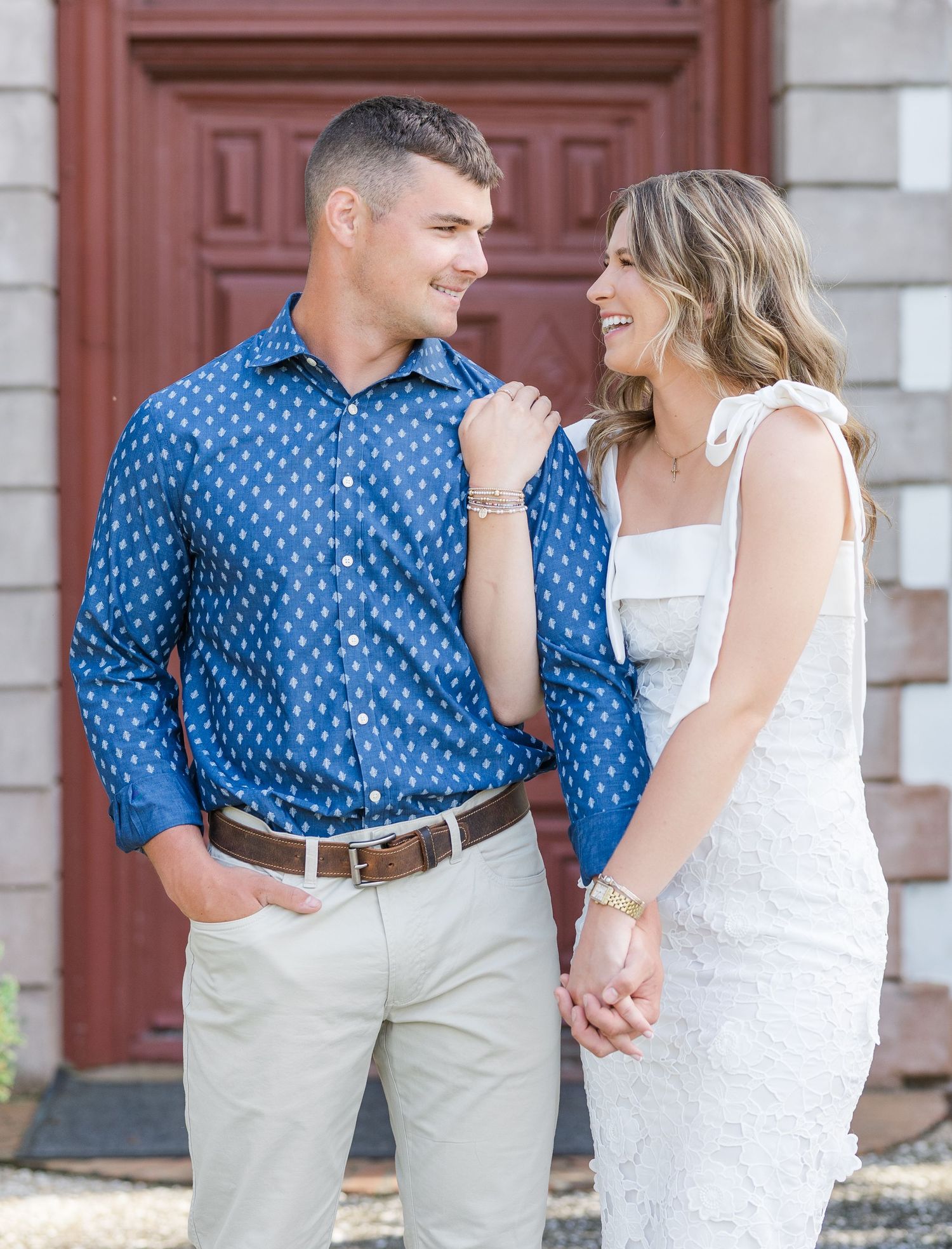 A smiling couple holds hands in front of a red door, man in blue patterned shirt and woman in white dress.