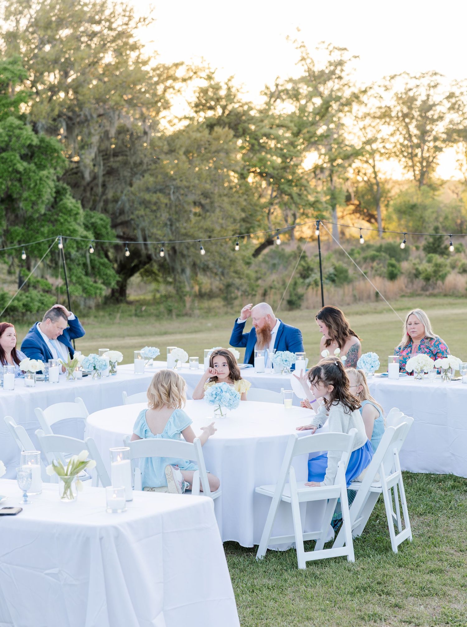 Guests seated at white-draped round tables during an outdoor wedding reception at sunset.