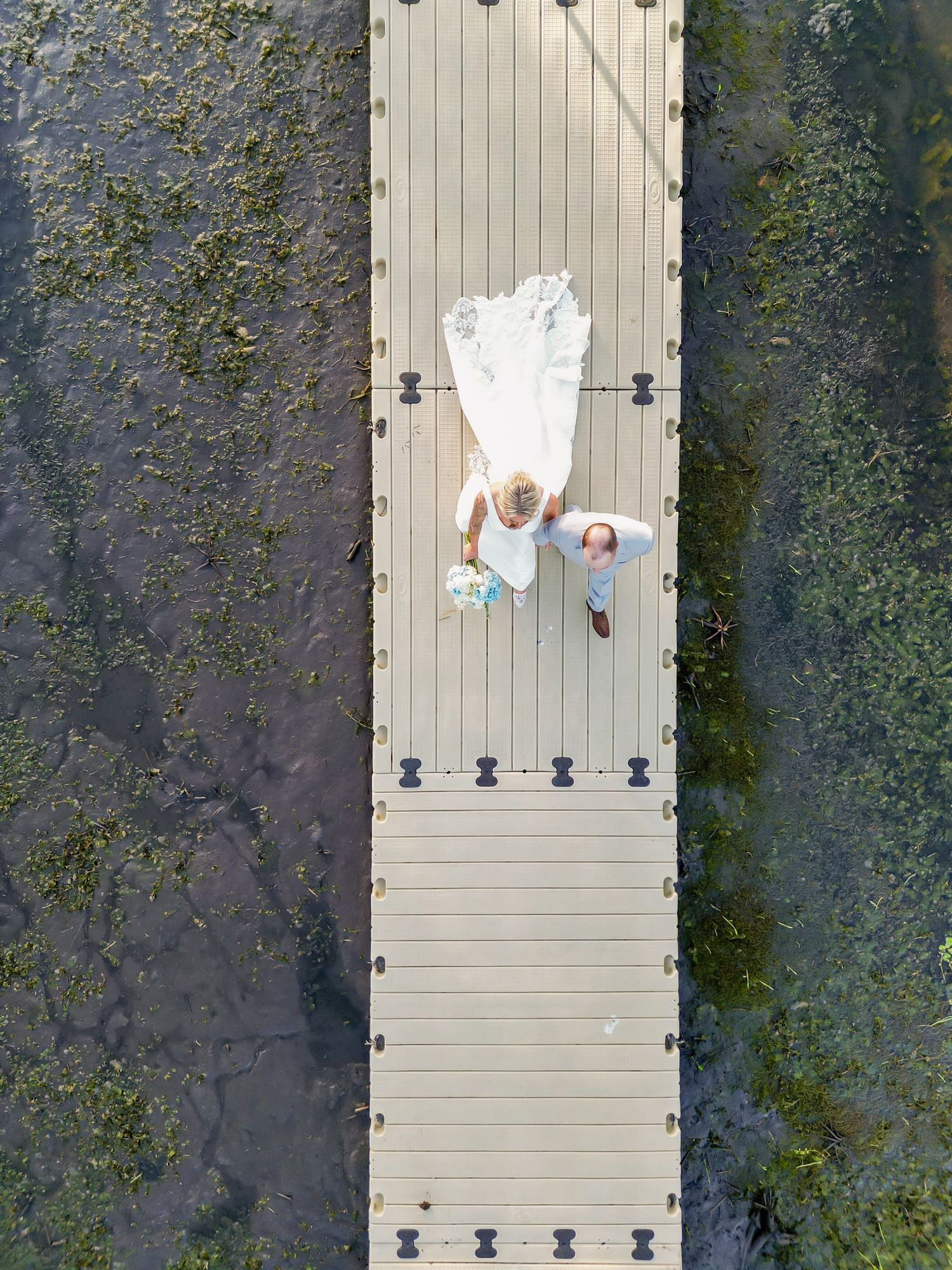 Aerial drone view of bride and groom lying on a wooden dock surrounded by green marsh water.