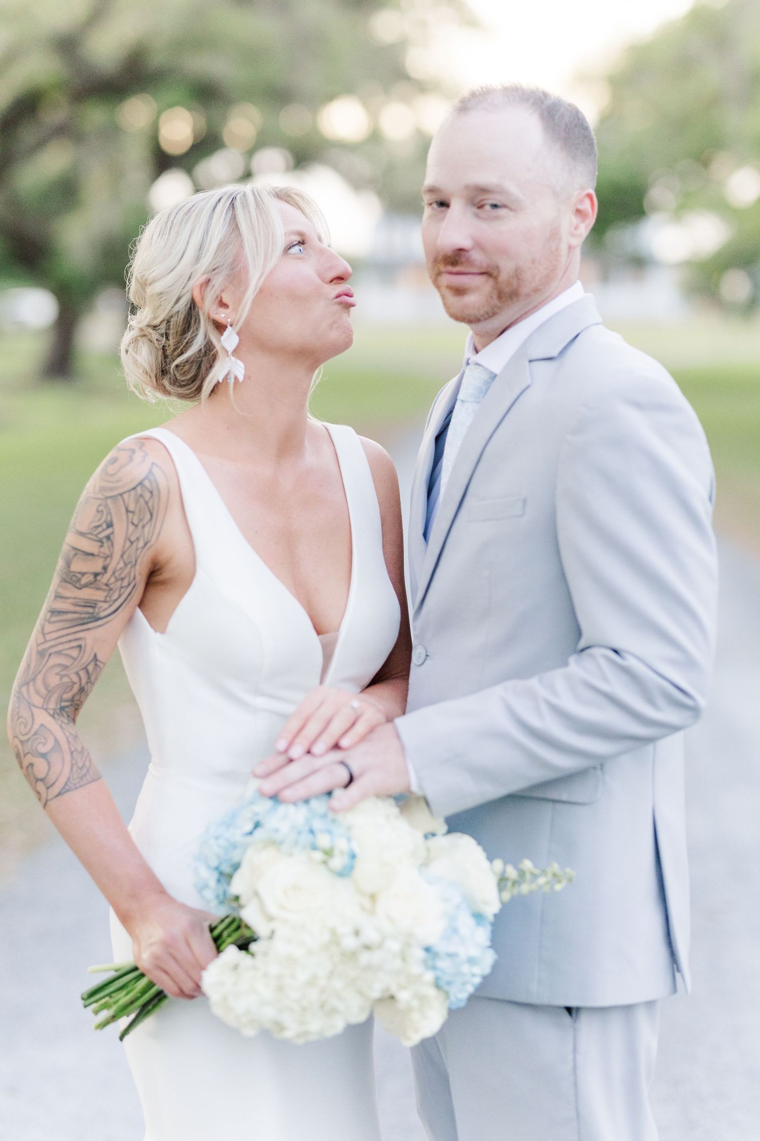 Bride with floral arm tattoo holding blue and white bouquet gazes at groom in gray suit outdoors.