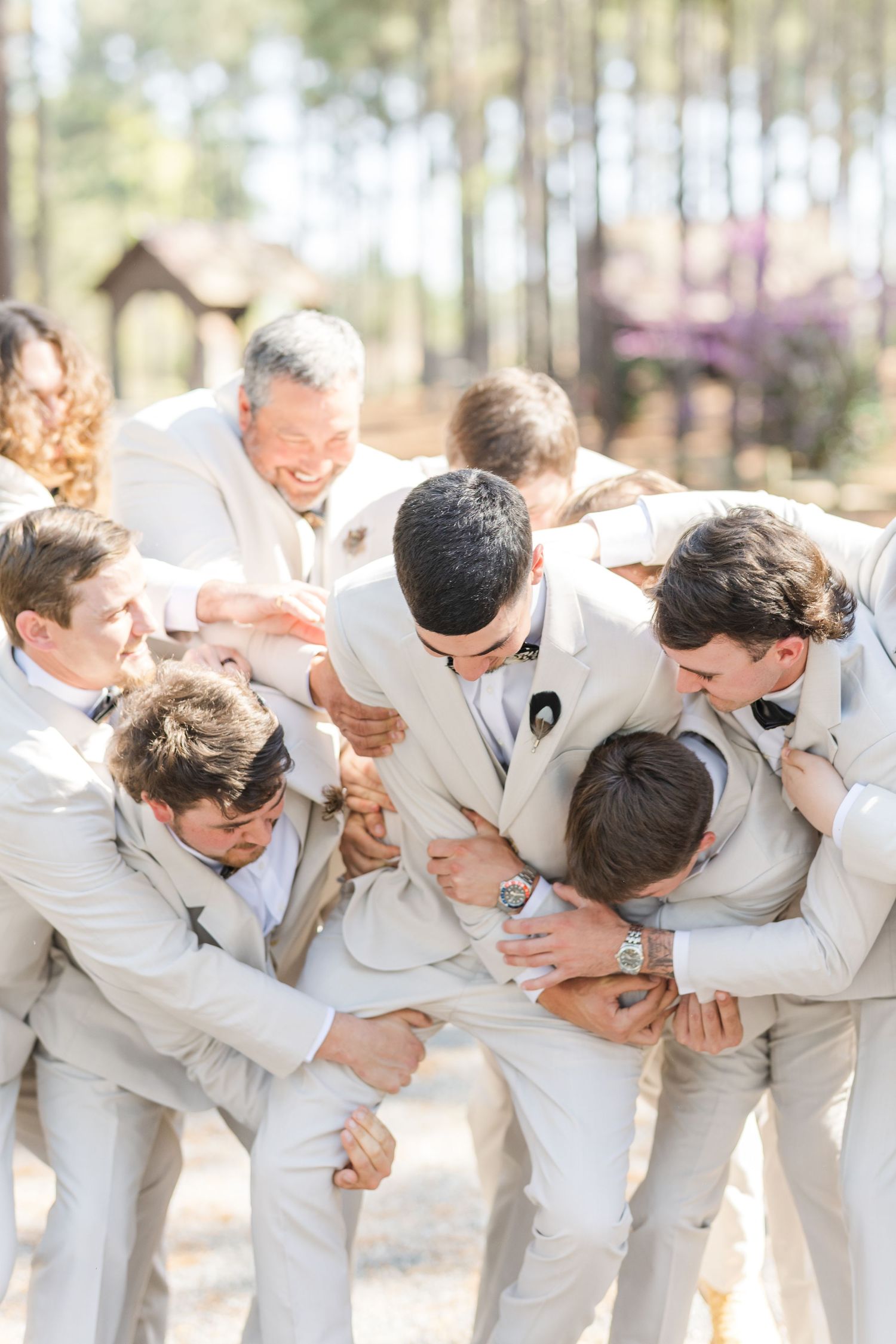 Groomsmen in tan suits playfully huddle around the groom outdoors in a joyful wedding celebration moment.