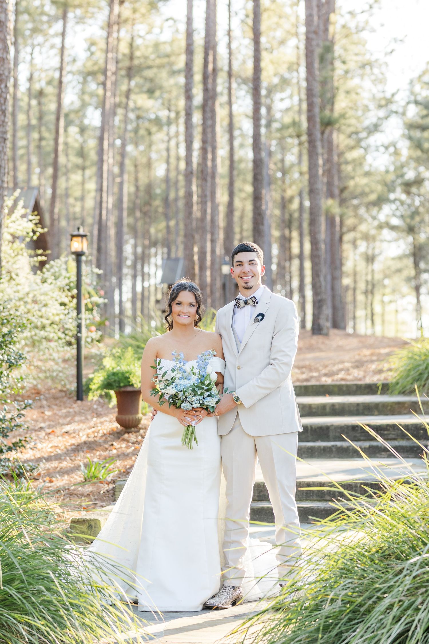 Bride and groom posing in a pine forest, groom in beige suit, bride holding blue bouquet.