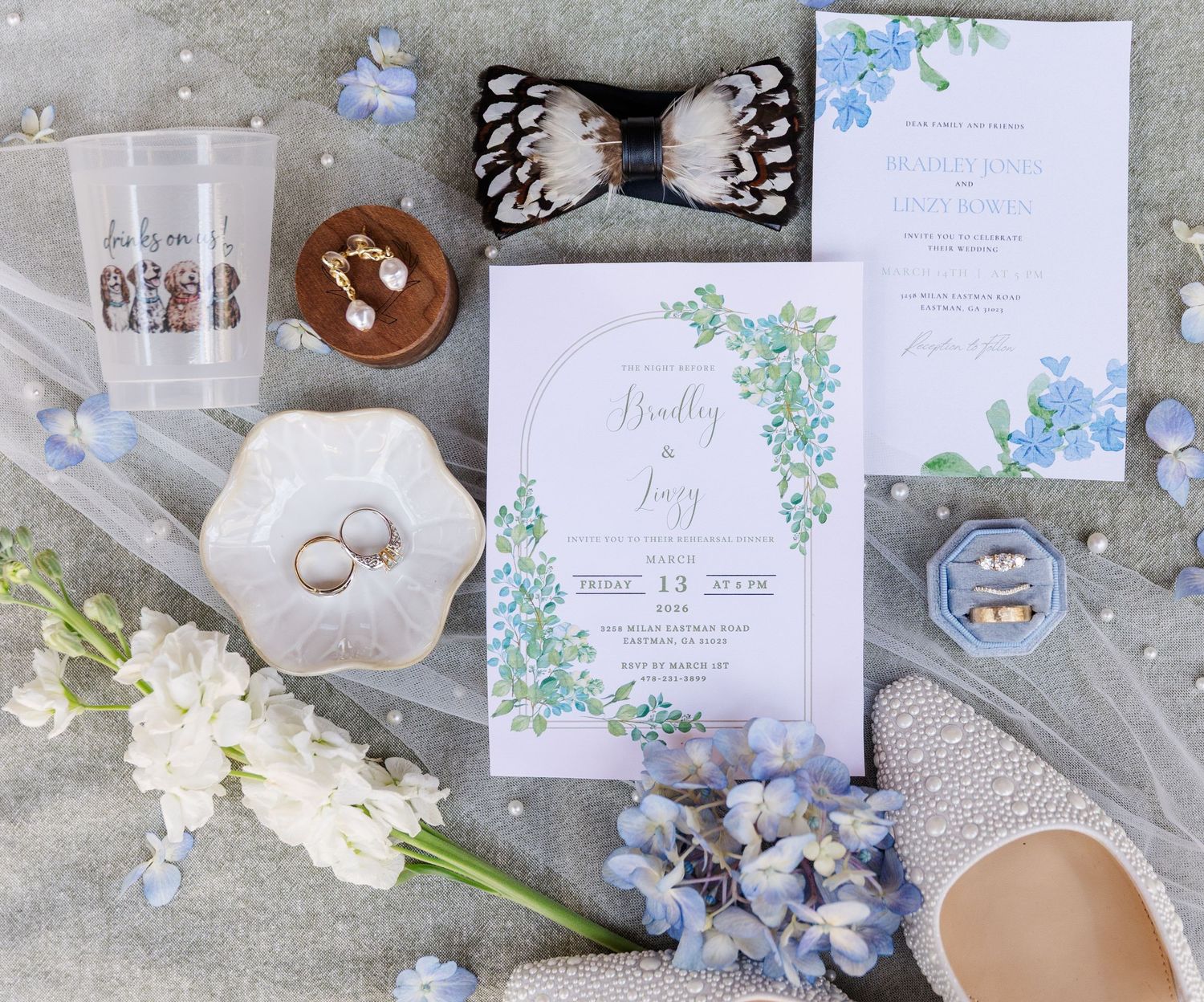 Wedding invitation suite flat lay with blue floral stationery, pearl heels, rings, bow tie, and delicate white and blue flowers.