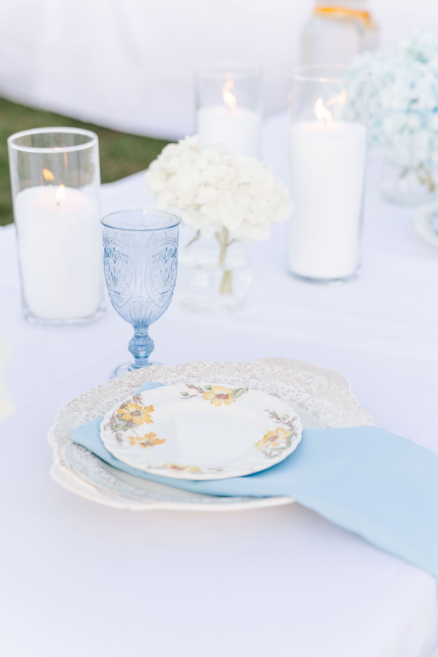 Romantic wedding table setting with blue goblet, floral china plates, lace doily, and white candles.