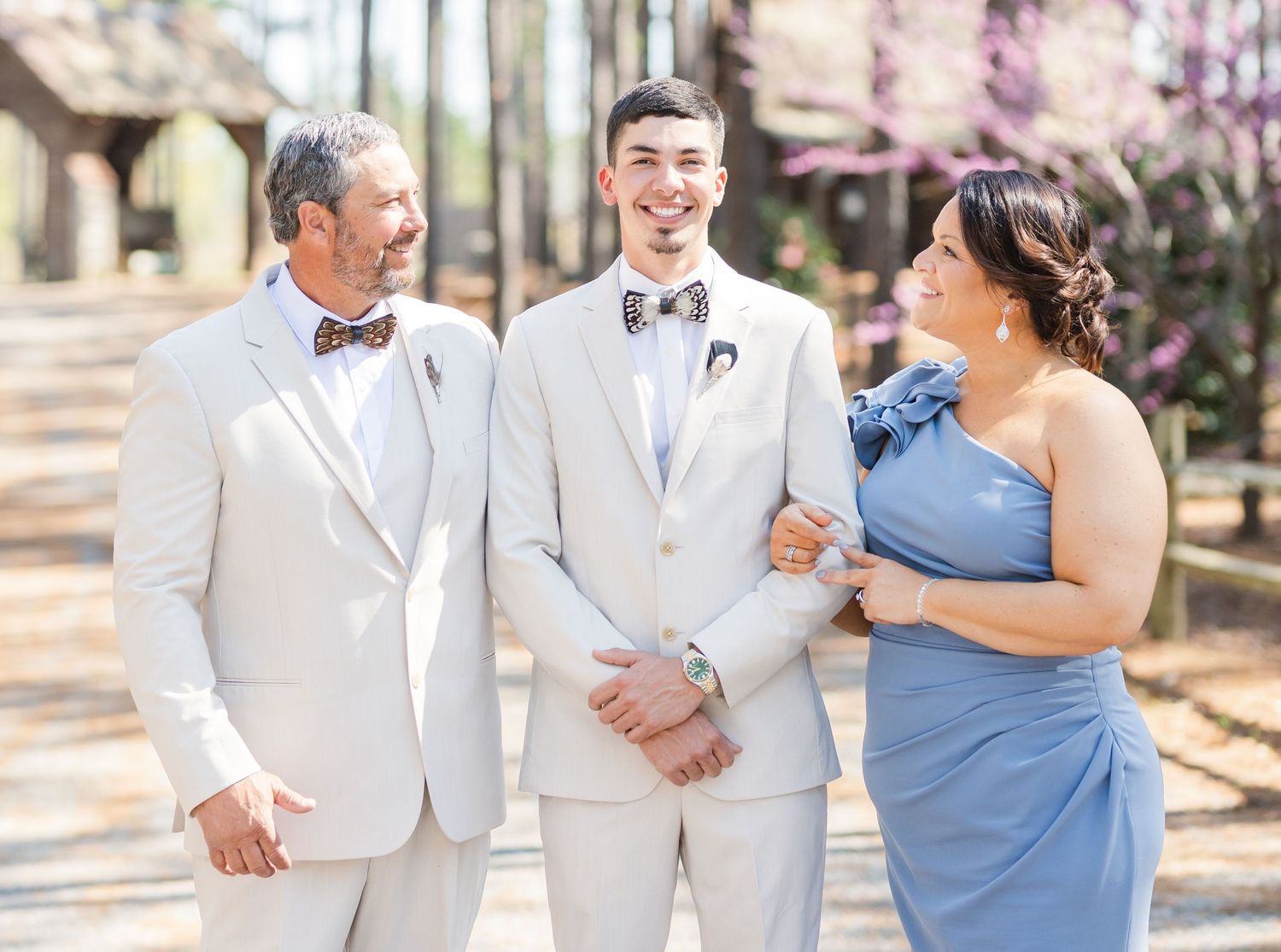Groom in cream suit smiling with parents wearing coordinating blue and tan formal attire outdoors.