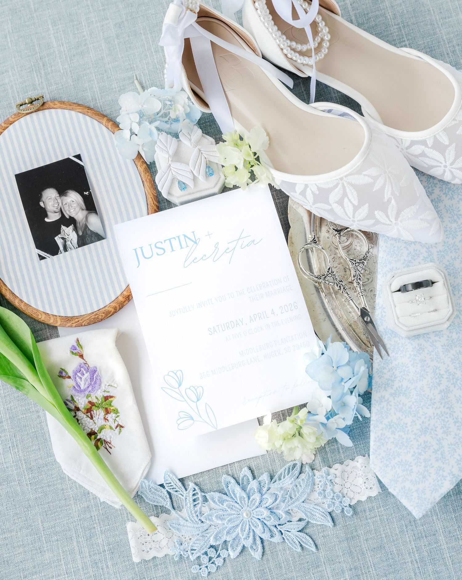 Elegant wedding flat lay with lace heels, floral invitation, blue flowers, ring box, and black-and-white photo on blue linen.