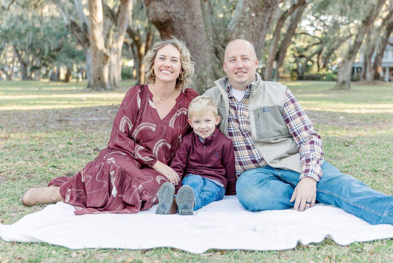 A family sits together on a white blanket under oak trees in a park setting.