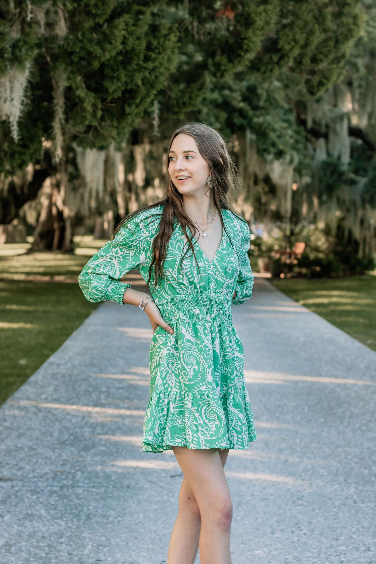 Woman in green patterned dress poses outdoors on path with Spanish moss trees.