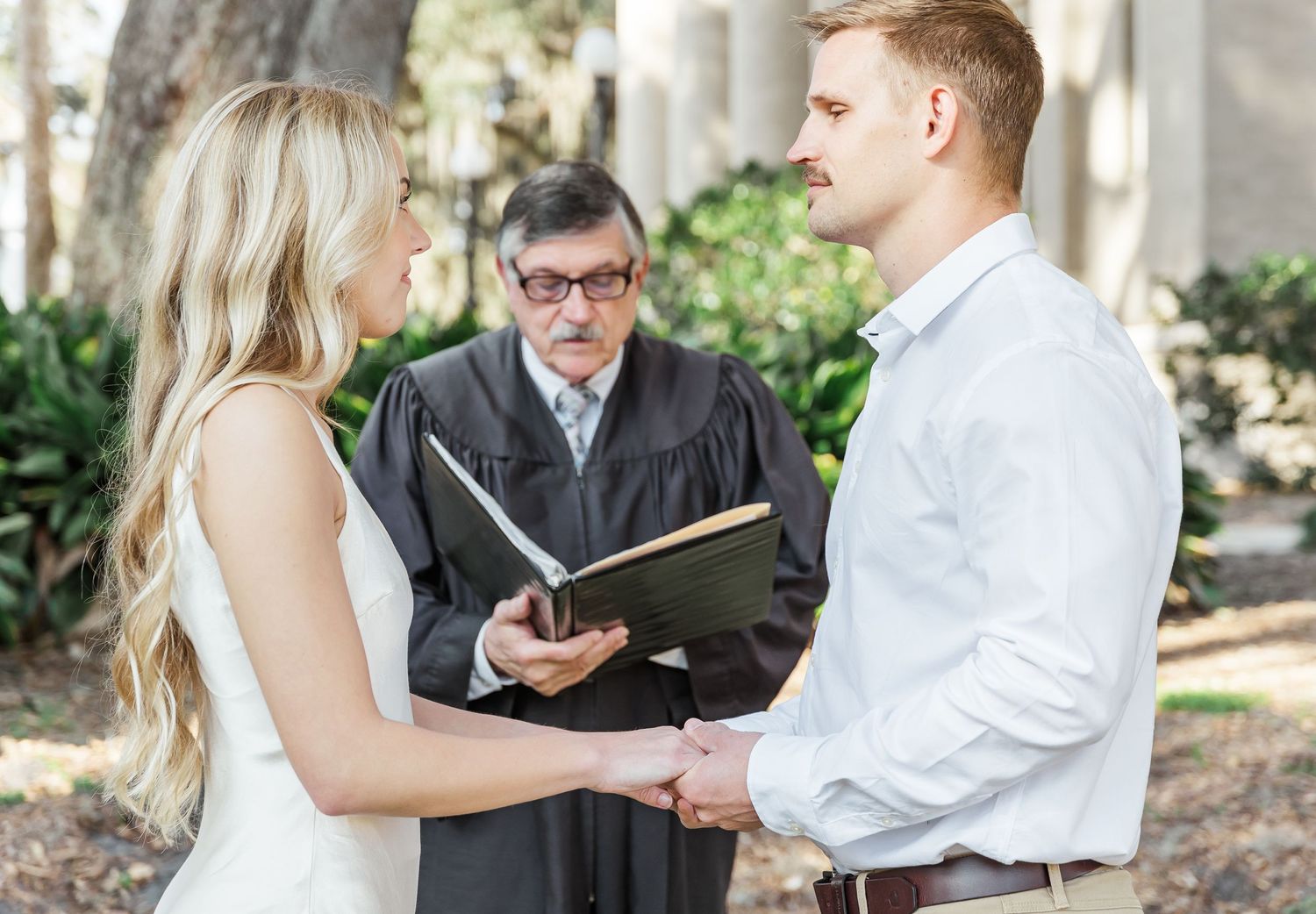 Outdoor wedding ceremony with officiant in black robes reading from book.