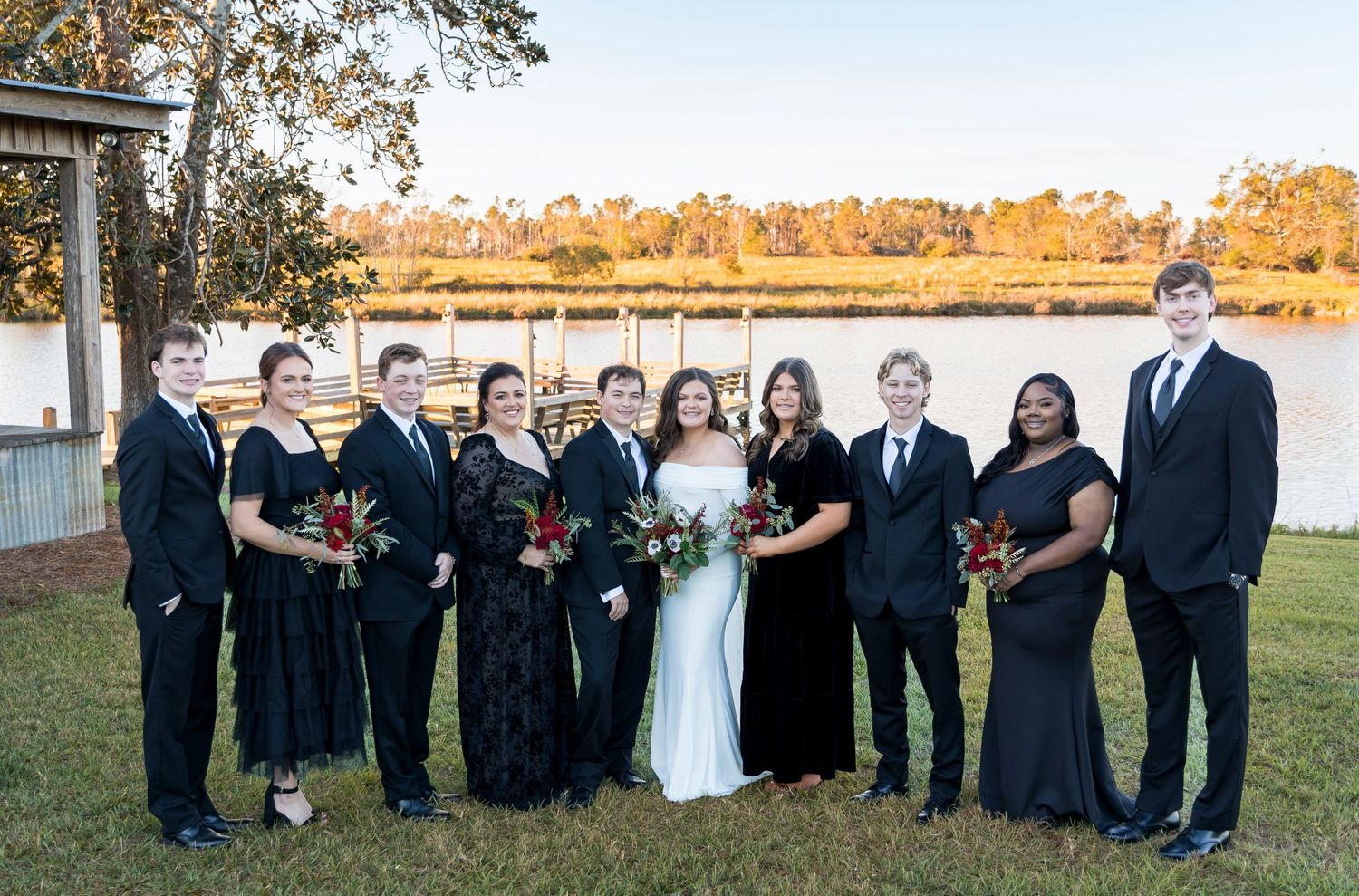 Wedding party in black attire and formal wear poses together by waterfront at sunset.