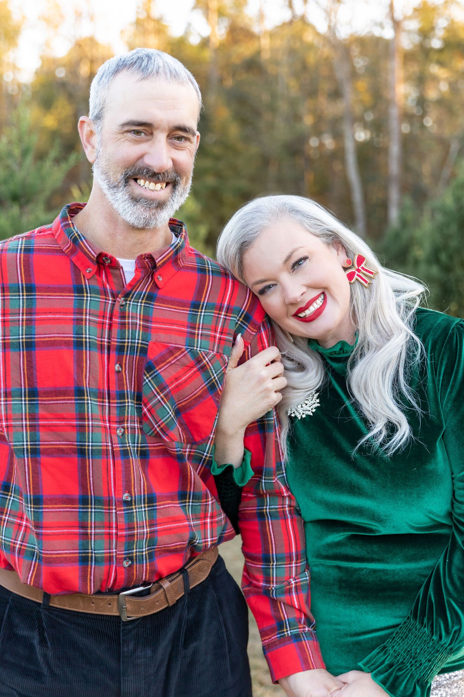 Couple in festive holiday attire with red plaid shirt and emerald velvet dress pose outdoors against golden sunset.