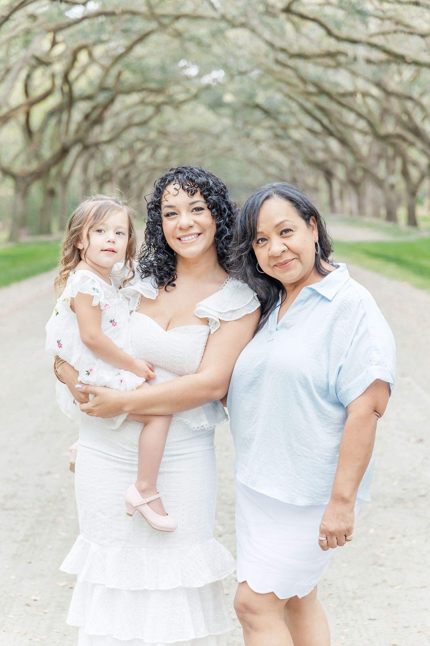 Three people in coordinating white and light blue outfits pose together on a scenic tree-lined path.