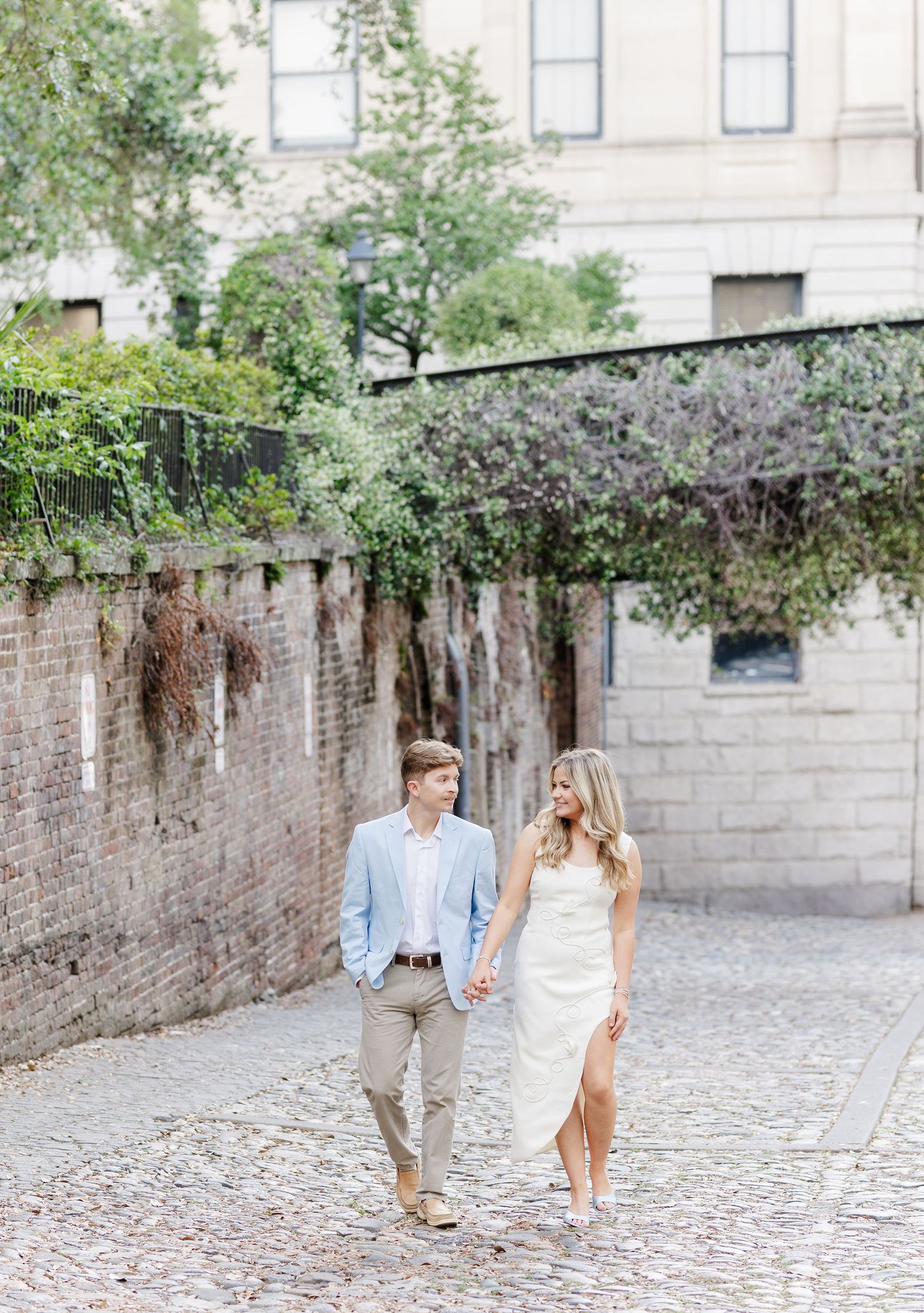 Couple holding hands walking on cobblestone street, man in light blue suit, woman in white dress, brick wall background.